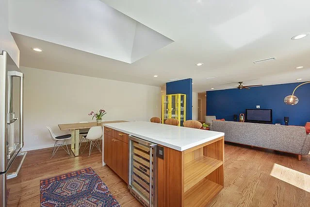 Open-concept living room and kitchen with white ceiling, hardwood floors, and modern furniture. The kitchen features a large white island with wooden cabinetry, a wine cooler, and a dining area with a white table and chairs. The living area has a gra