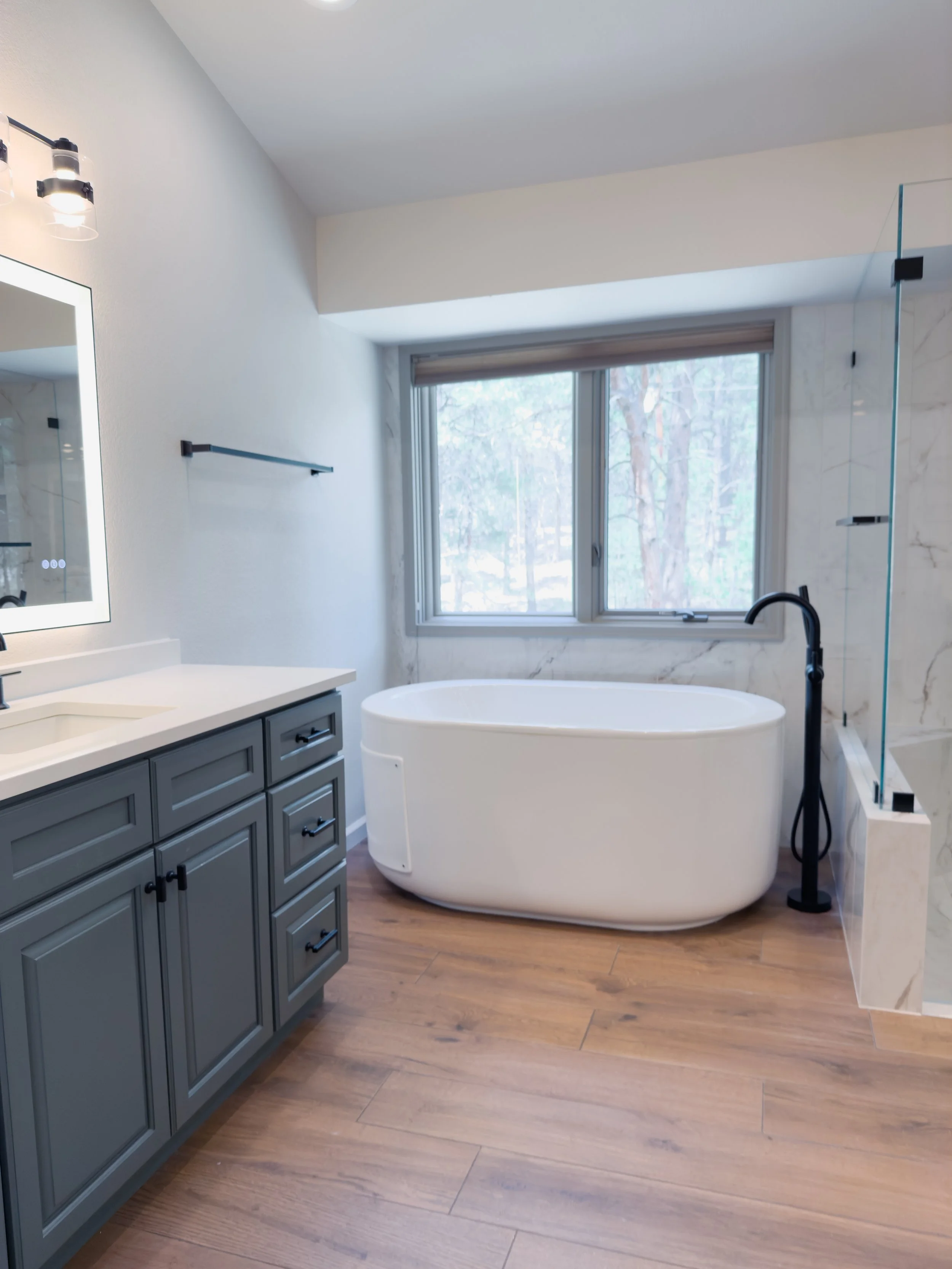 Modern bathroom with a white freestanding bathtub near a window, gray vanity with black hardware, wooden flooring, and a glass-enclosed shower.