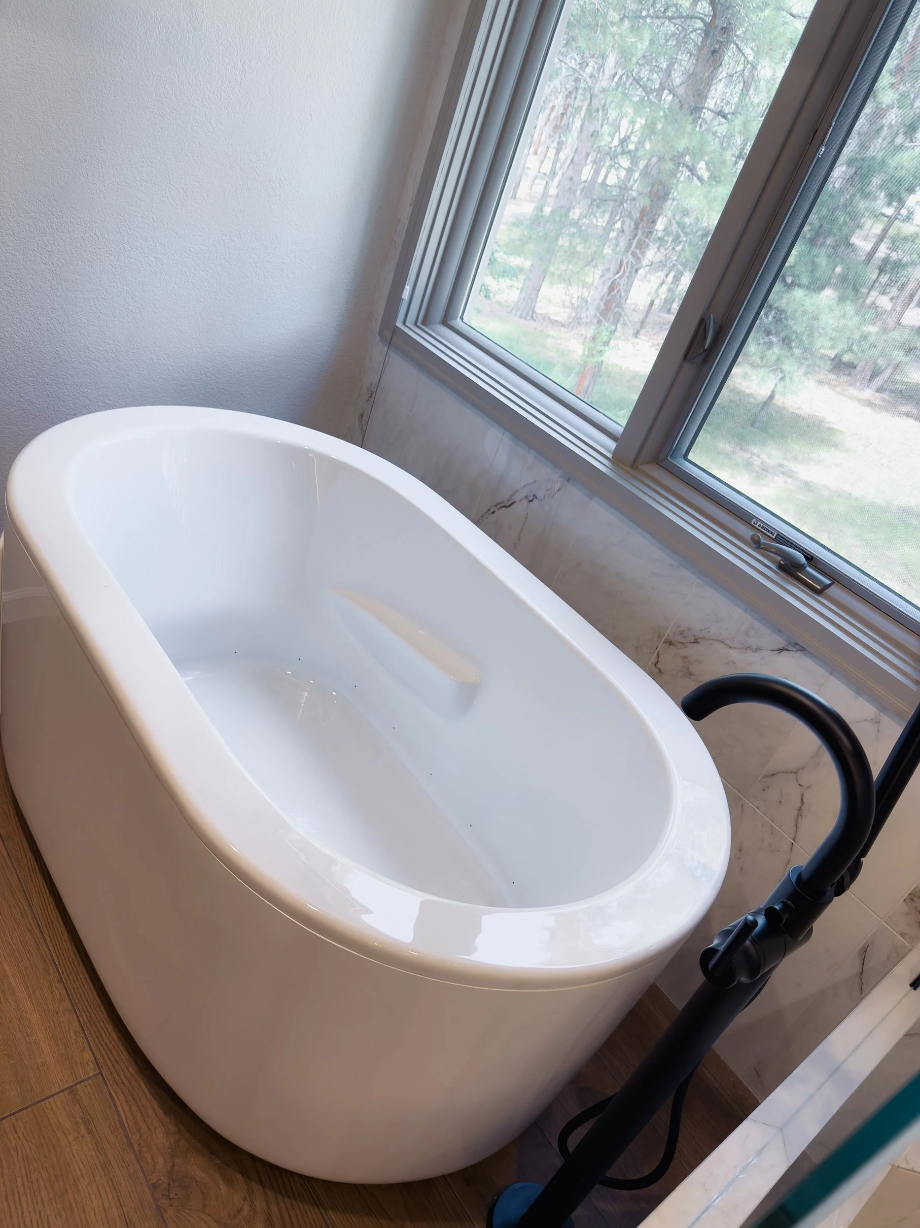 White freestanding bathtub next to a window with forest view, black floor-mounted faucet, and brown wood flooring.