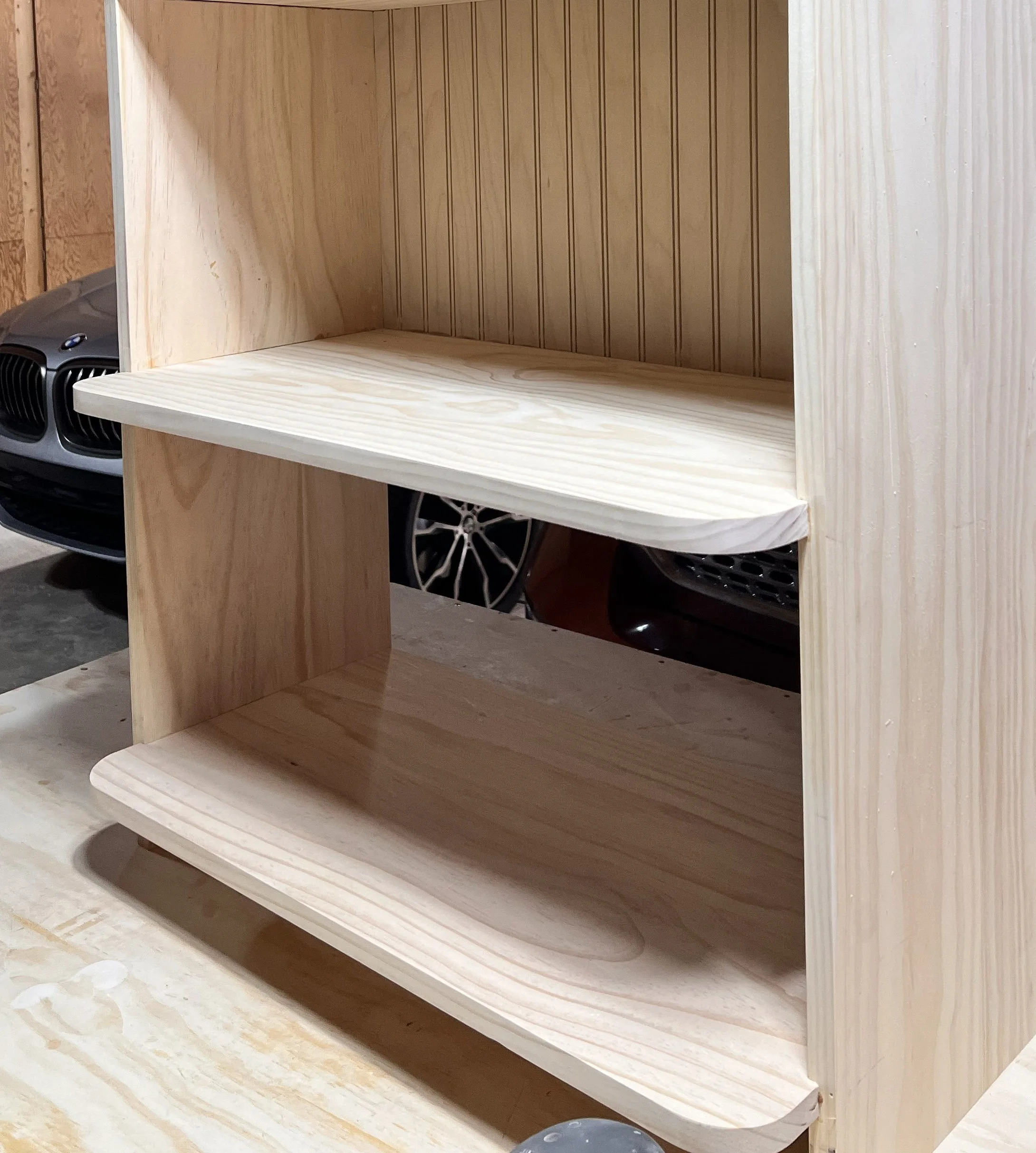 Unfinished wooden shelf with two tiers, made of light-colored wood, sitting on a workbench in a workshop.