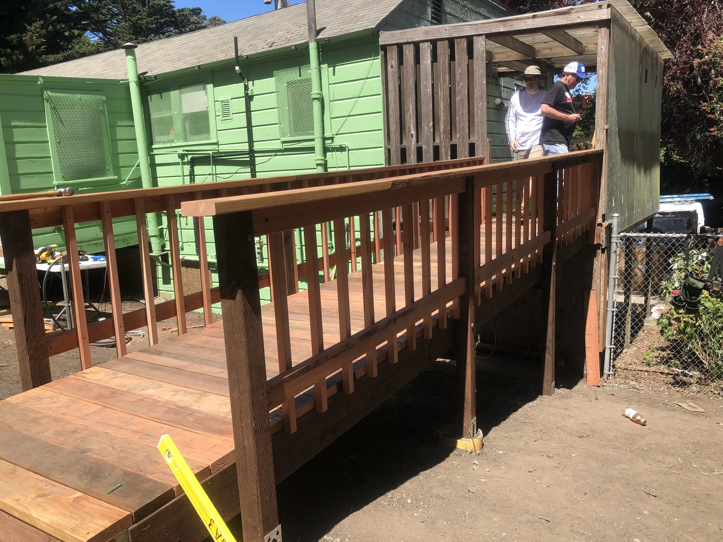 Two people working on a wooden deck extension outside a green building, with tools and construction materials around.