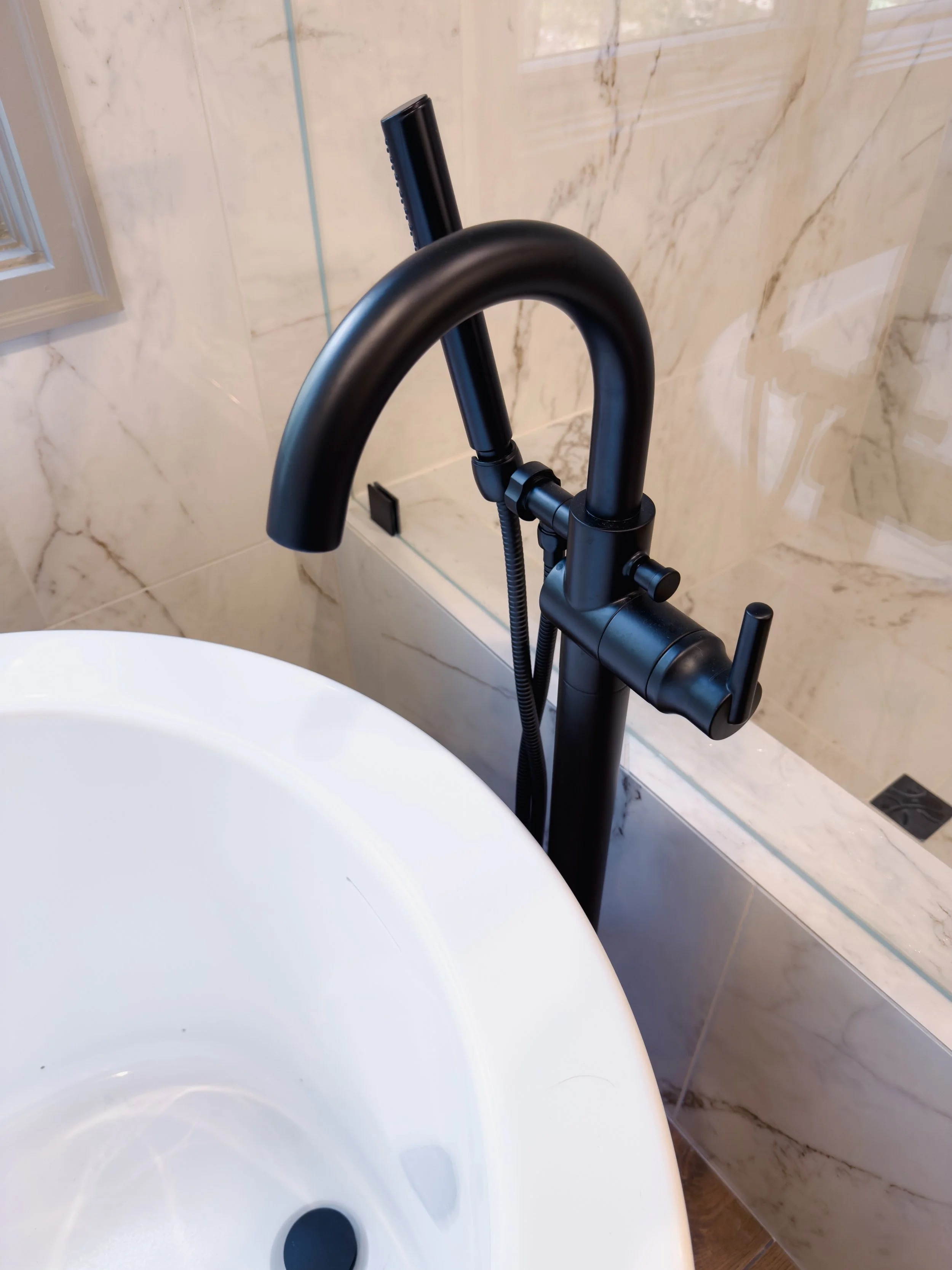 A black bathroom faucet with a black handheld showerhead attached, mounted next to a white sink in a marble-tiled bathroom.
