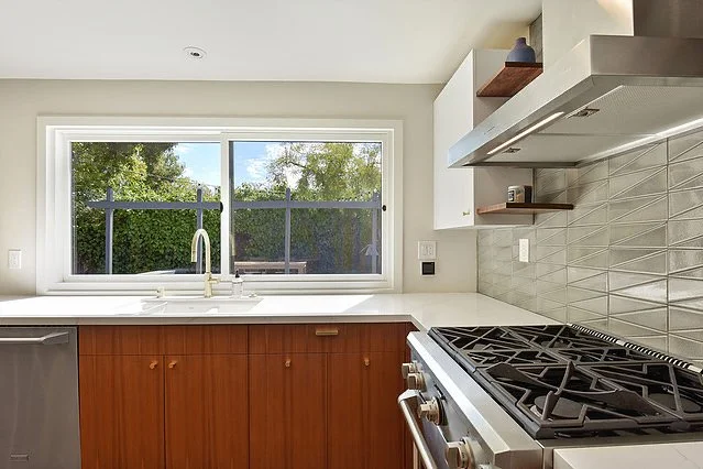 Modern kitchen with a window above the sink, white cabinets, wooden lower cabinets, stainless steel stove, and a tiled backsplash.