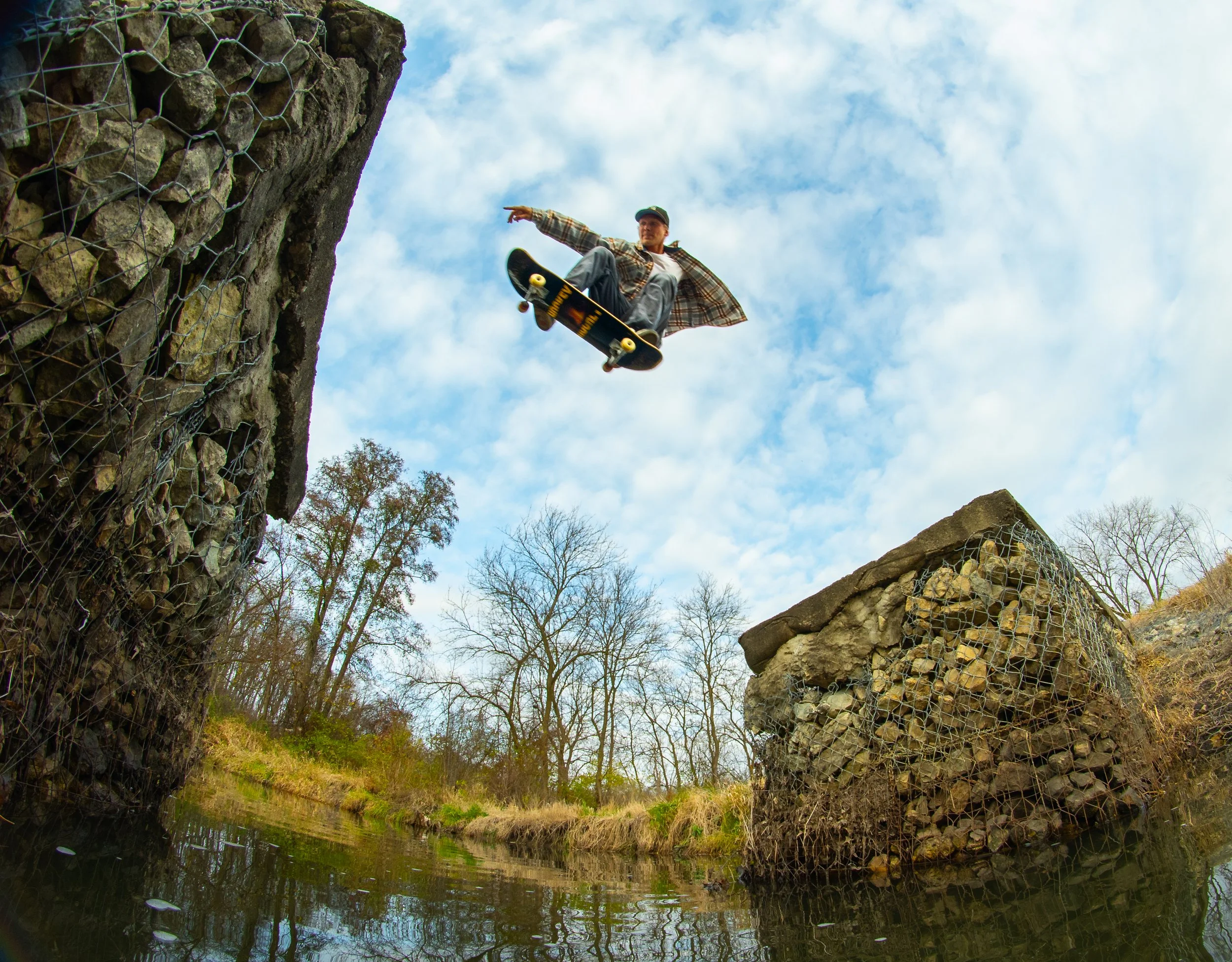 Skateboarder in mid-air performing a trick over a small body of water with stone and wire structures on either side and leafless trees in the background under a partly cloudy sky.