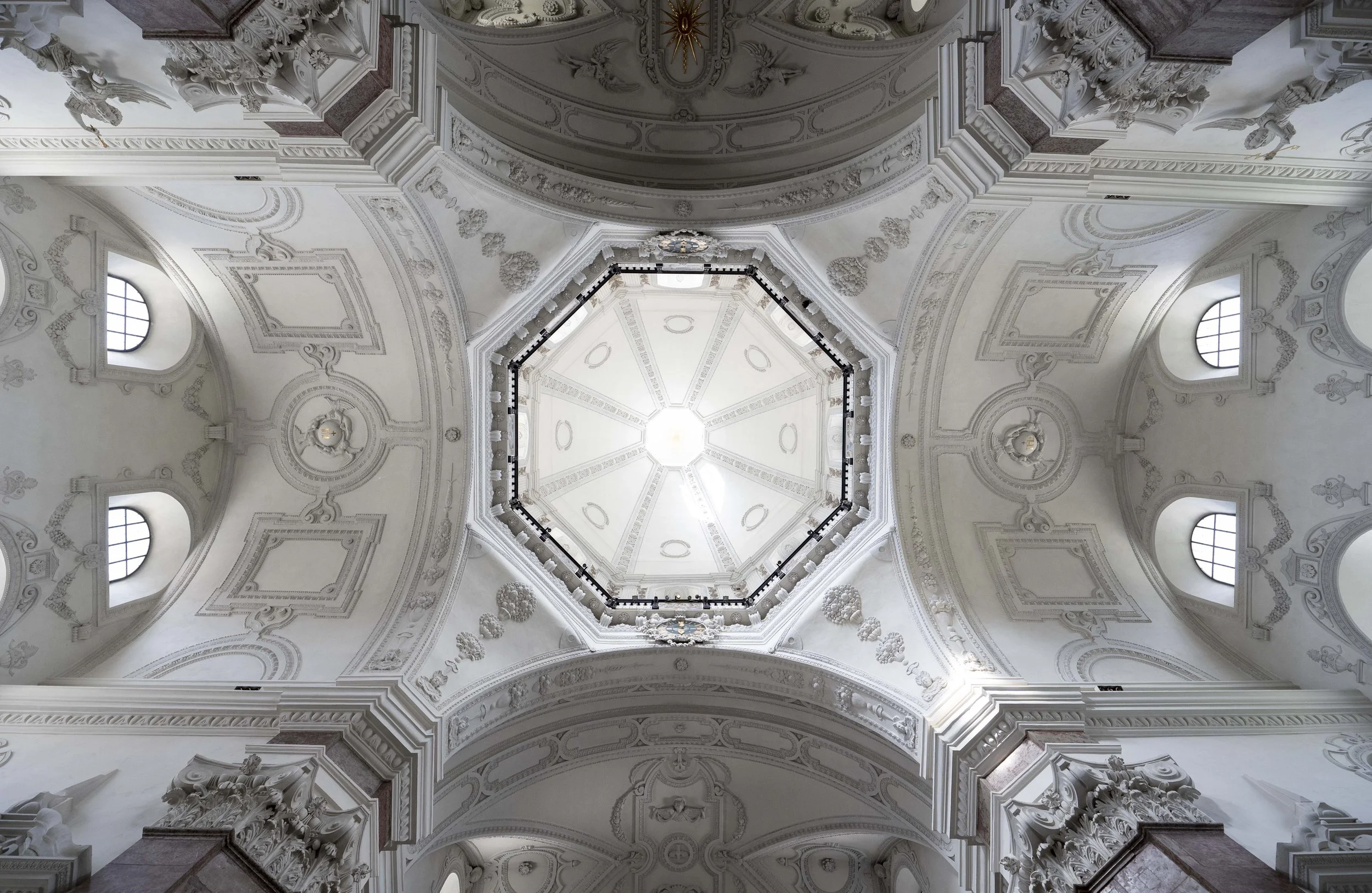 View of a decorated, white domed ceiling inside a church or cathedral, with windows and ornate architectural details.