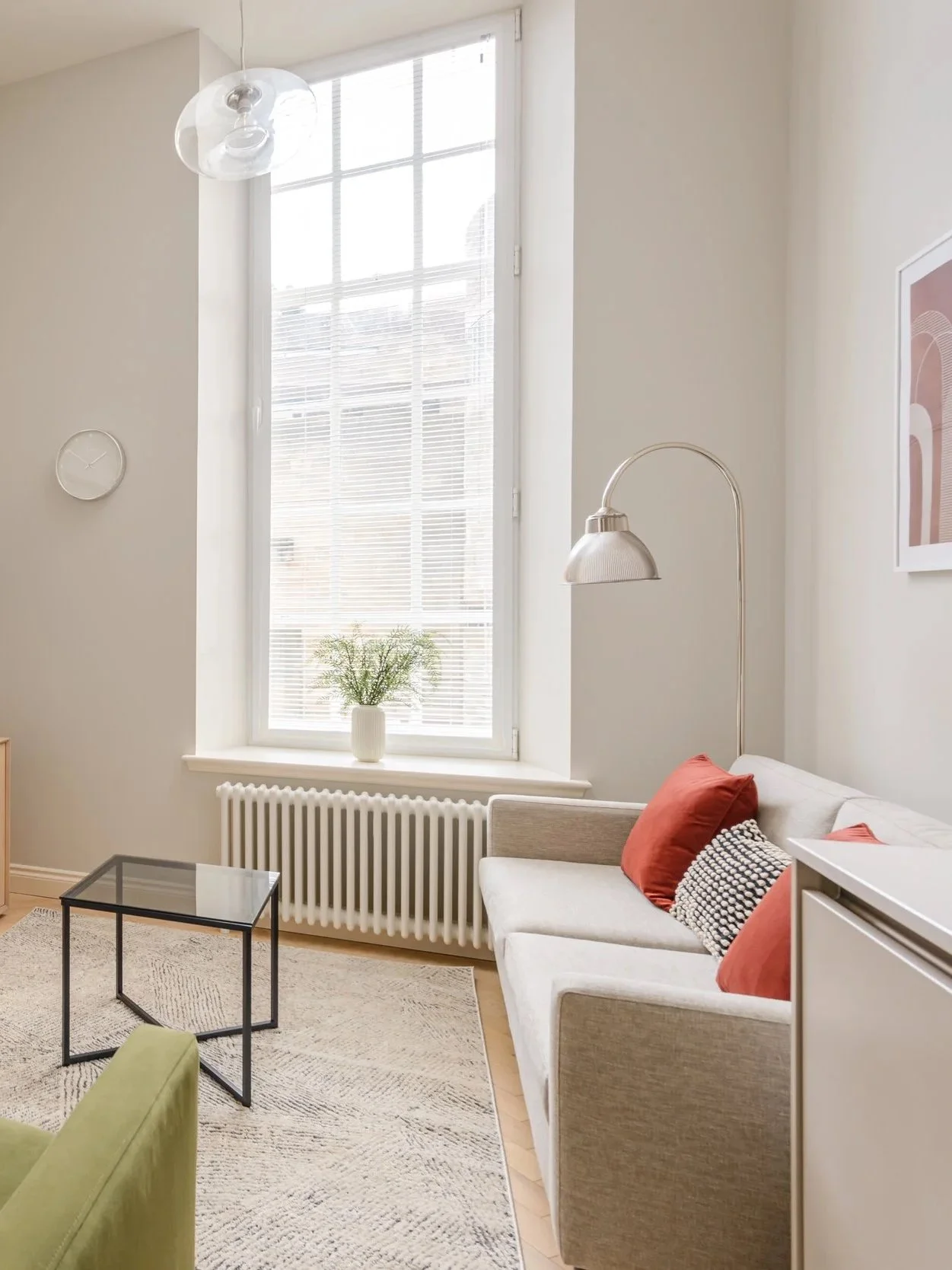 A living room with a large window, a white sofa with red and black pillows, a small glass-top coffee table, a green chair, and a tall arched floor lamp. There's a white vase with greenery on the window sill, a textured rug, a wall clock, and framed artwork.
