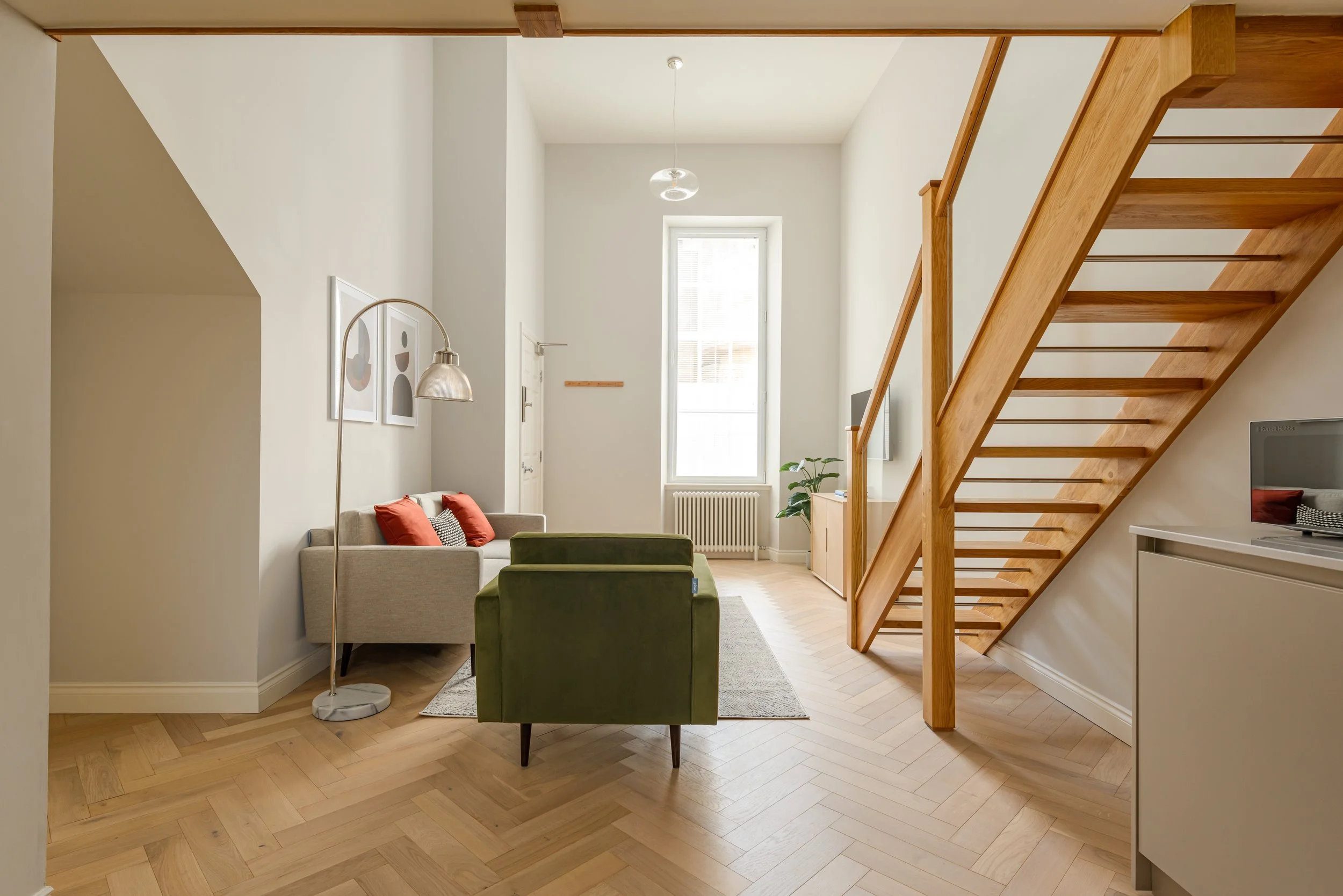 Bright living room with high ceiling, large window, wooden staircase, beige sofa with red cushions, green armchair, floor lamp, artwork on wall, and light-colored wooden flooring.