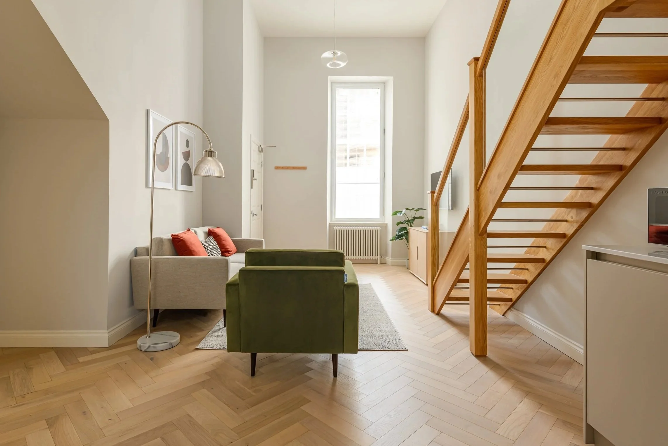 Modern living room with white walls, wooden staircase, and large window. Includes a beige sofa with red pillows, a green armchair, floor lamp, and framed artwork on the wall.
