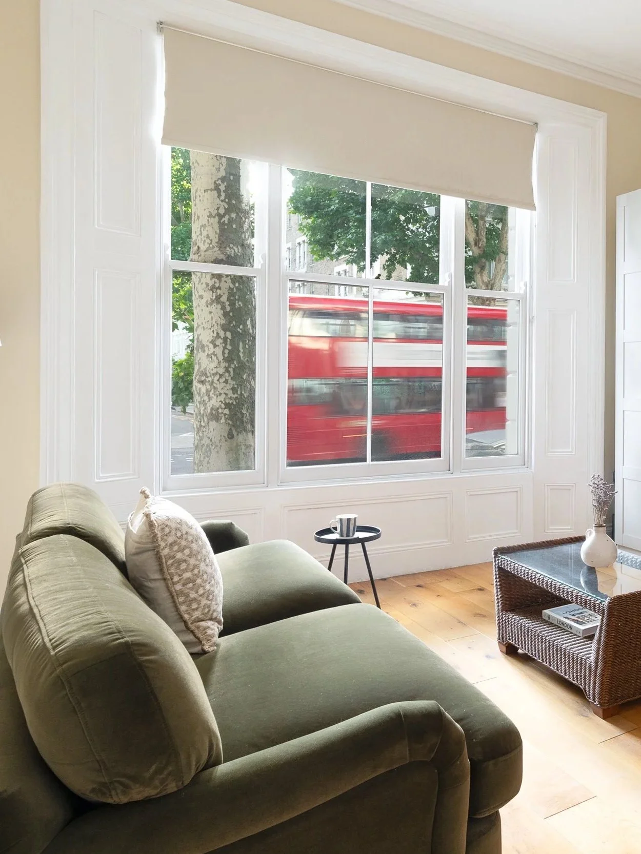 Living room with green couch, decorative pillows, small round side table with mug, wicker coffee table with books and vase, large window with white trim and beige roller shade, view of trees and red bus outside.