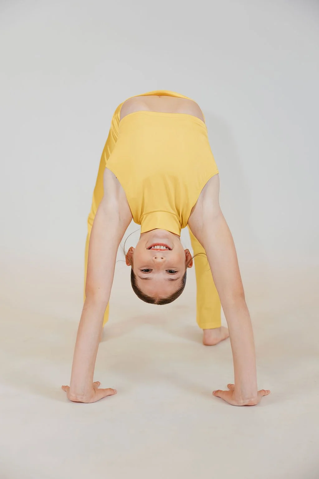 A young girl with brown hair in a ponytail is performing a yoga pose, bending upside down with her hands and feet on the ground, wearing a yellow outfit, and smiling at the camera.