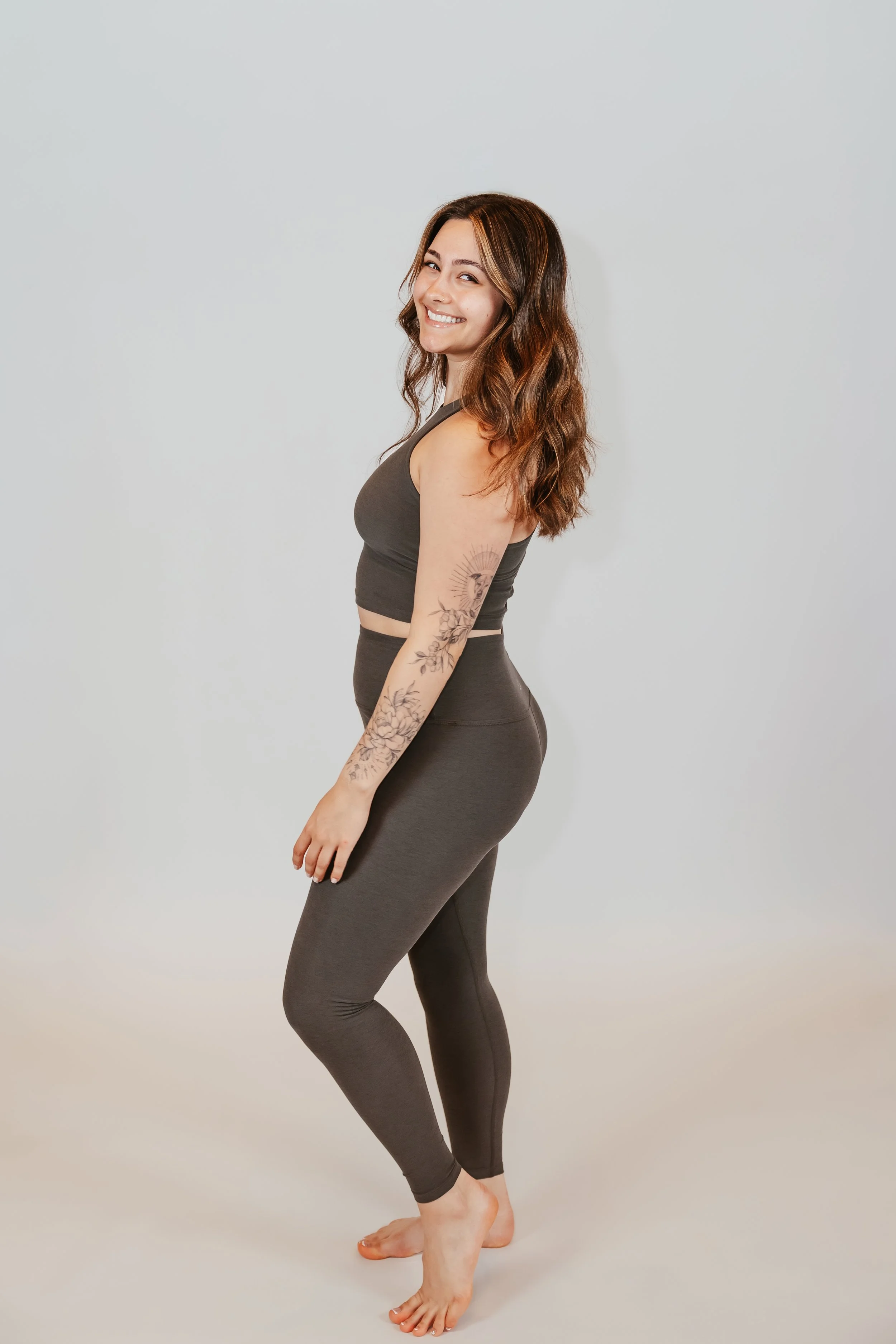 A woman with long wavy brown hair, tattoo on her left forearm, smiling and standing on her toes barefoot against a light gray background.