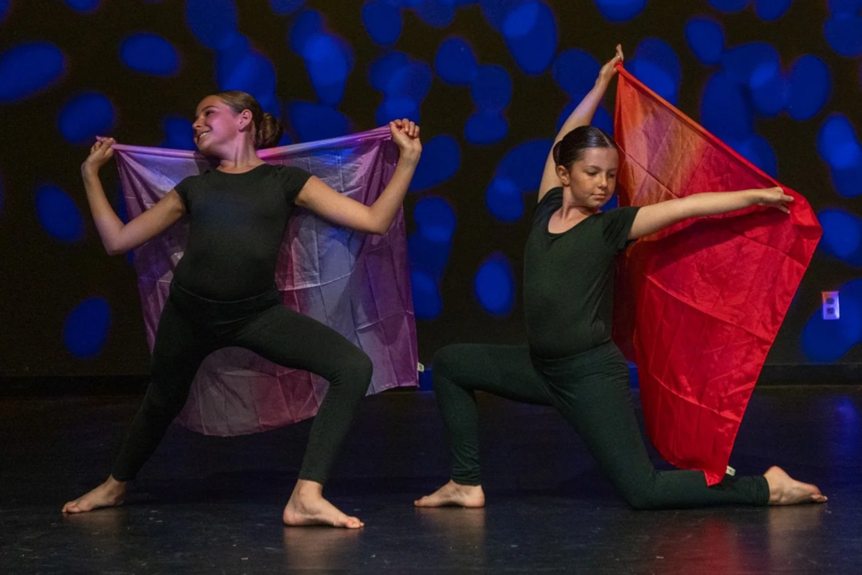 Two young women perform a dance with large, colorful scarves on a stage with a dark background illuminated by blue lights.