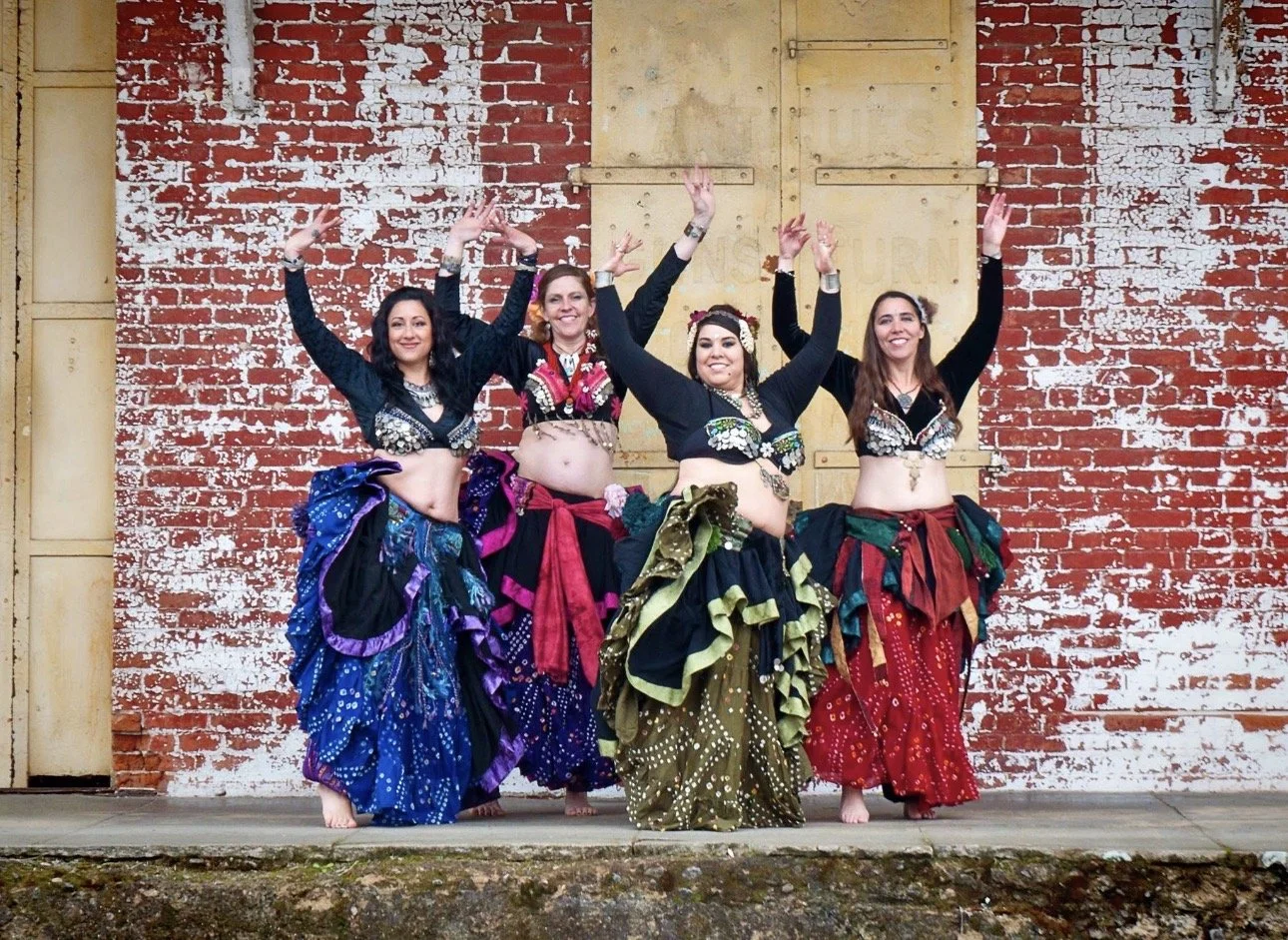Four women dancing in colorful belly dance costumes with colorful skirts and black tops, posing with hands raised, standing in front of a rustic brick wall.