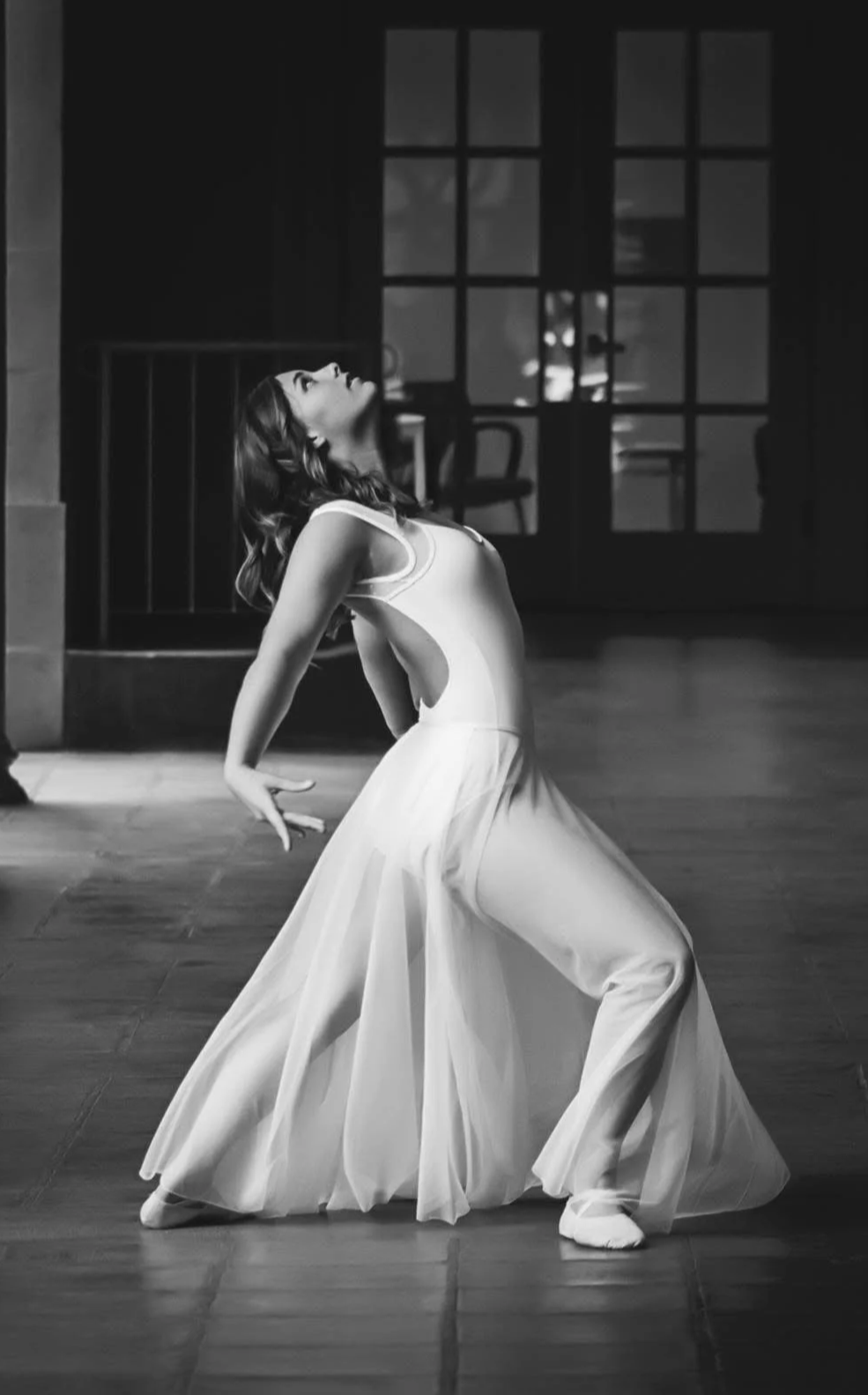 A woman in a flowing white dress dancing on a wooden floor in a dimly lit room, with a large window with multiple panes in the background.