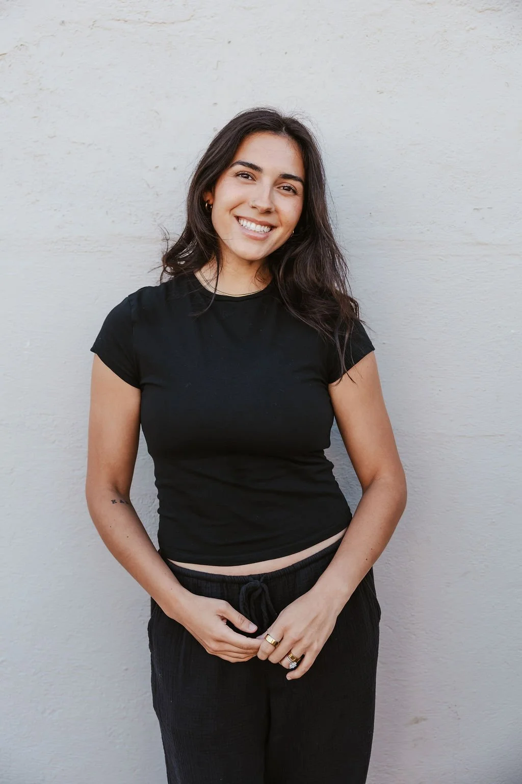A young woman with long dark hair, smiling, wearing a black t-shirt and black pants, standing against a plain white wall.