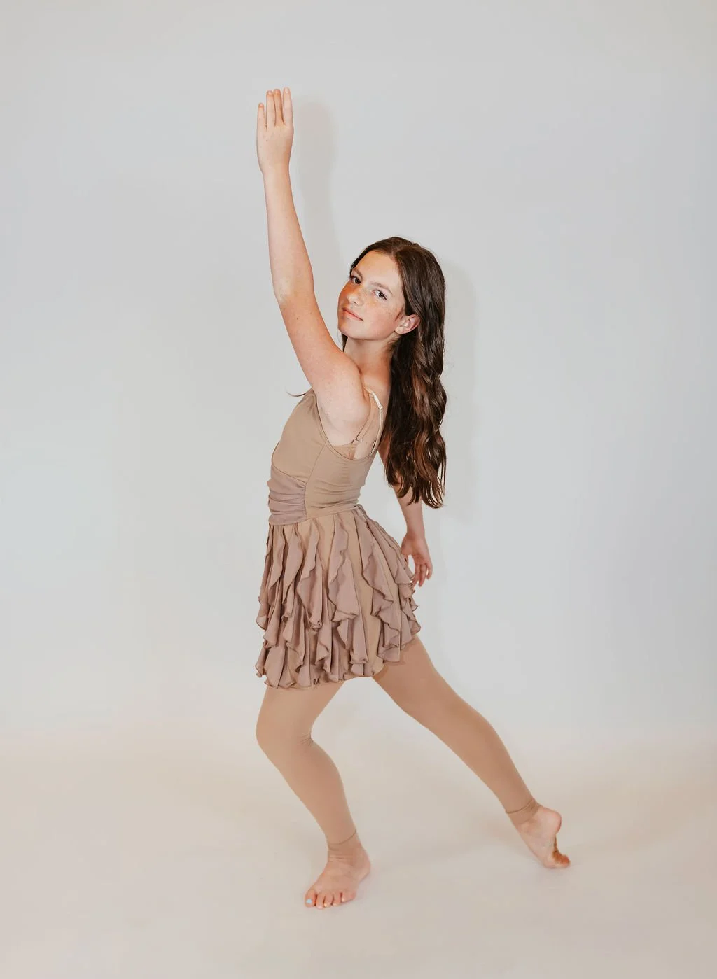 A young girl with long brown hair wearing a beige dance costume with a ruffled skirt, posing against a plain white background.