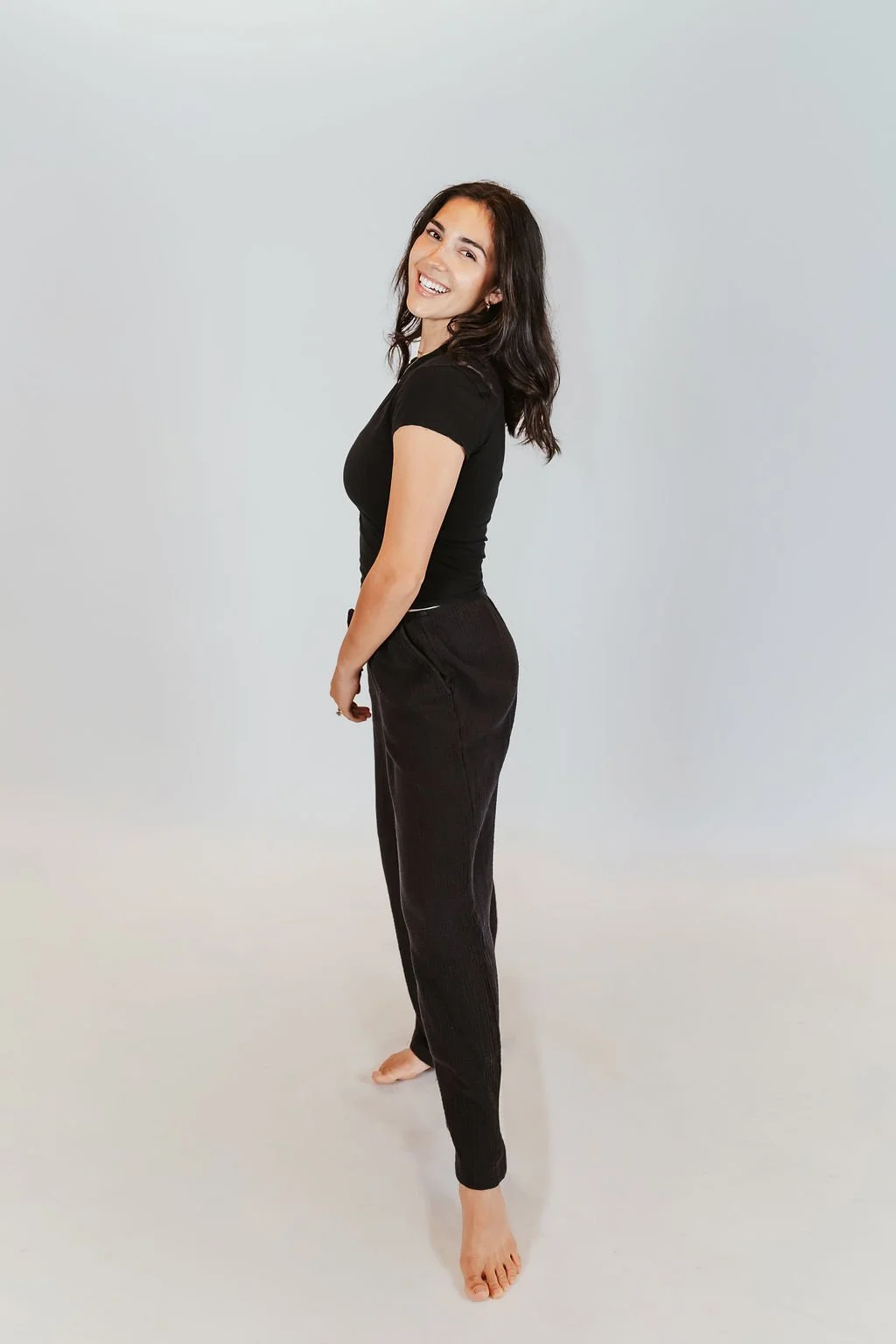 A woman with long dark hair, wearing a black t-shirt and black pants, standing barefoot against a plain white background, smiling and looking at the camera.