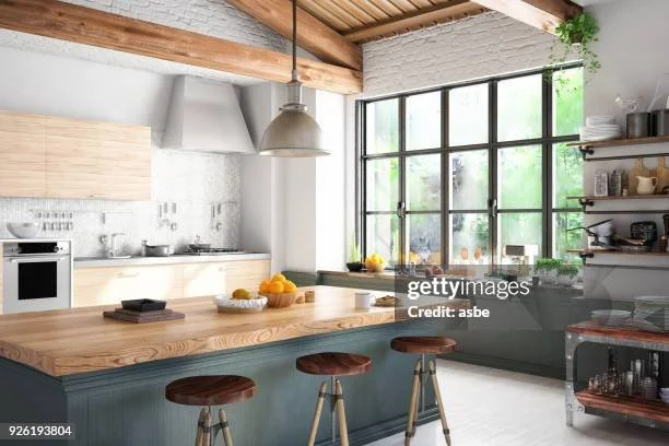 Modern kitchen with wooden ceiling beams, large window, wooden island with three stools, and open shelving with dishes and plants.