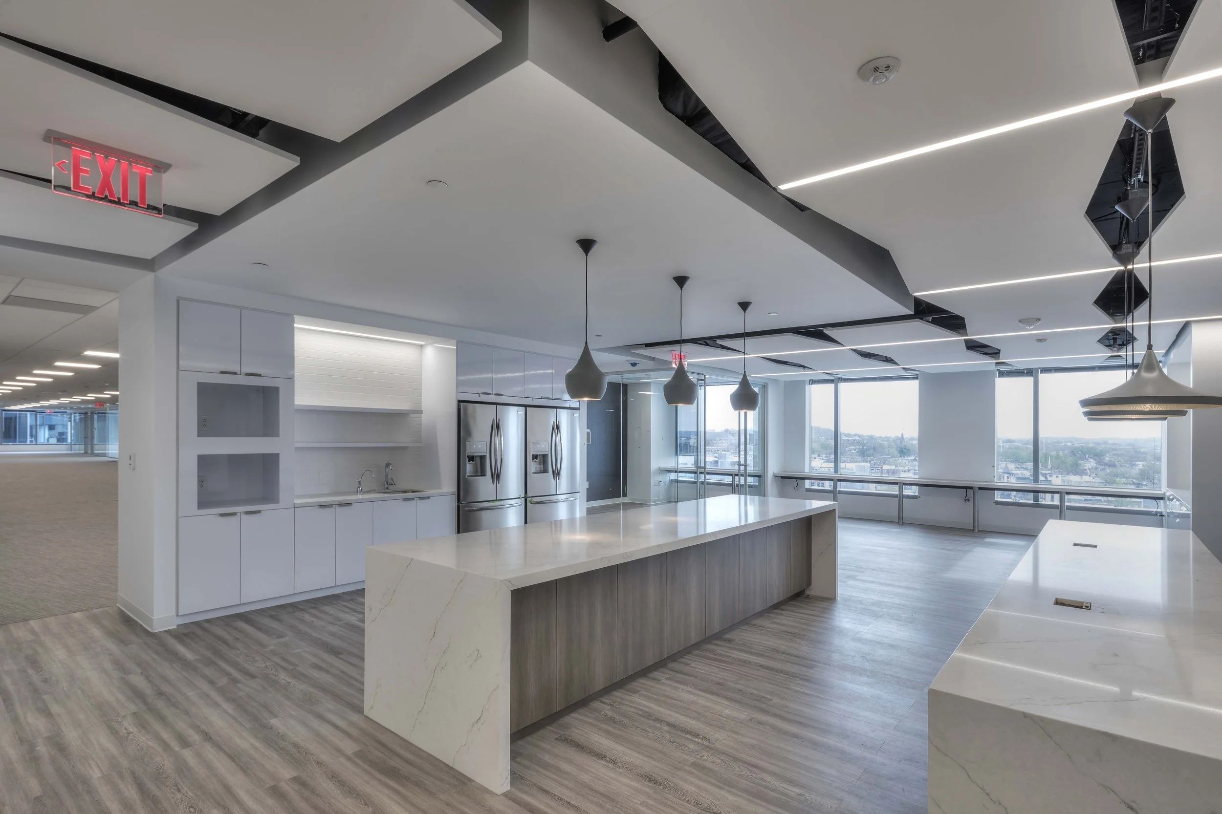 Modern, empty kitchen area with white and wood finishes, large central island, stainless steel refrigerators, and large windows with city view.