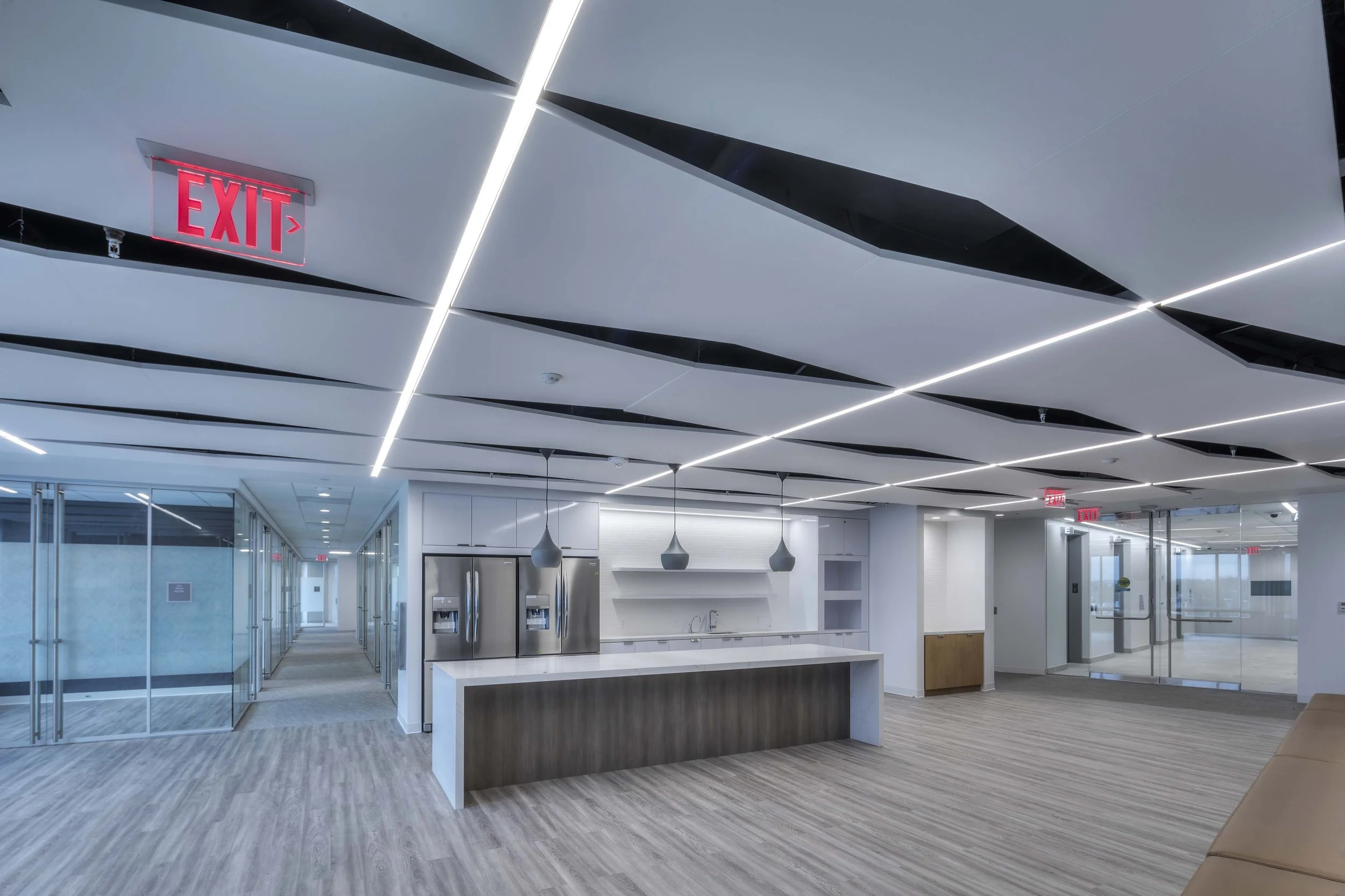 Modern office break room with a kitchen island, two stainless steel refrigerators, pendant lights, and glass walls.