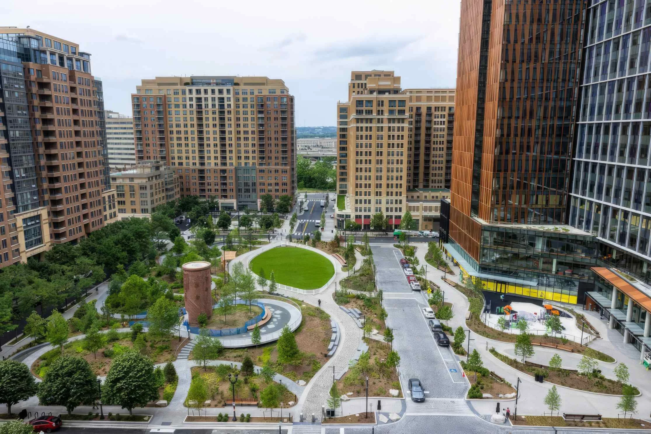 A view of a city park surrounded by tall modern buildings with a green lawn, trees, walking paths, and parked cars.
