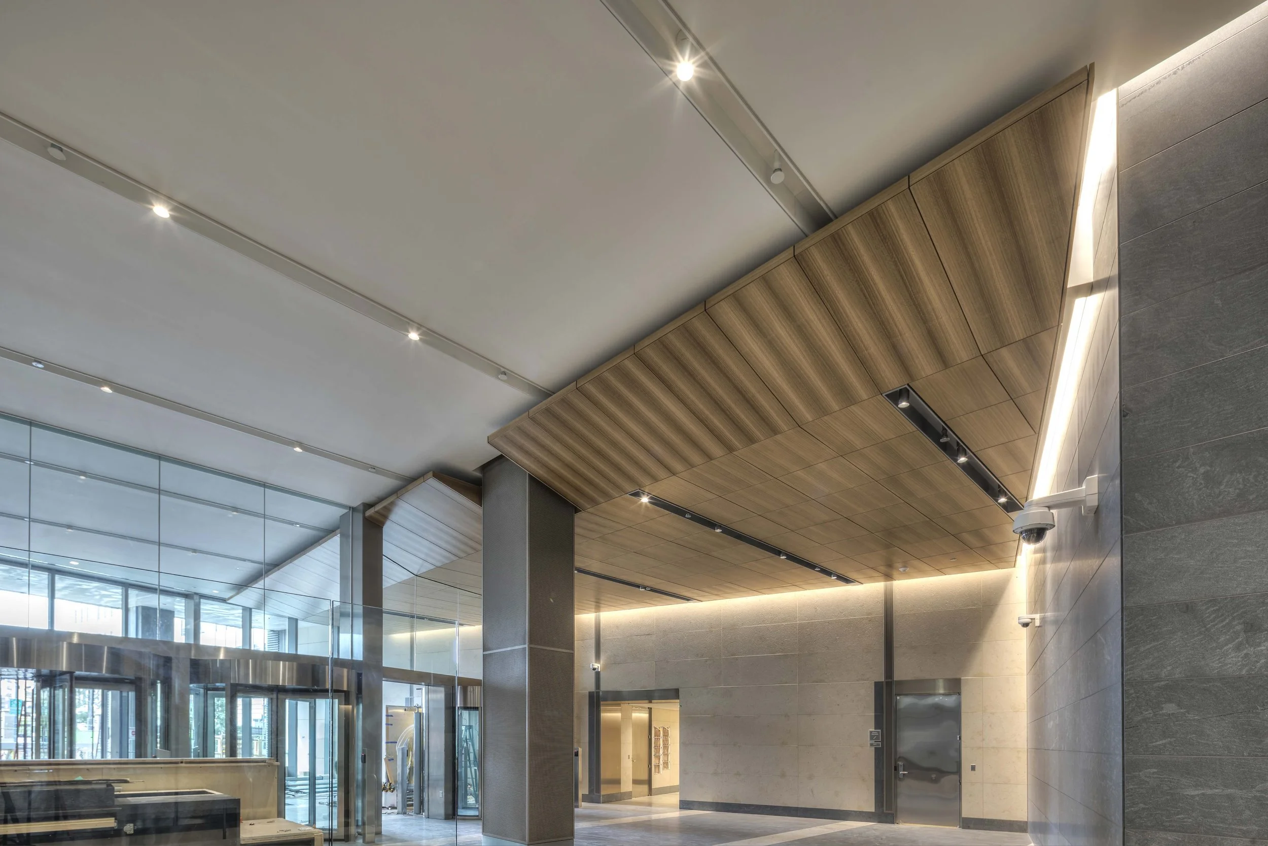Modern building lobby with glass walls, wooden ceiling panels, and security cameras.