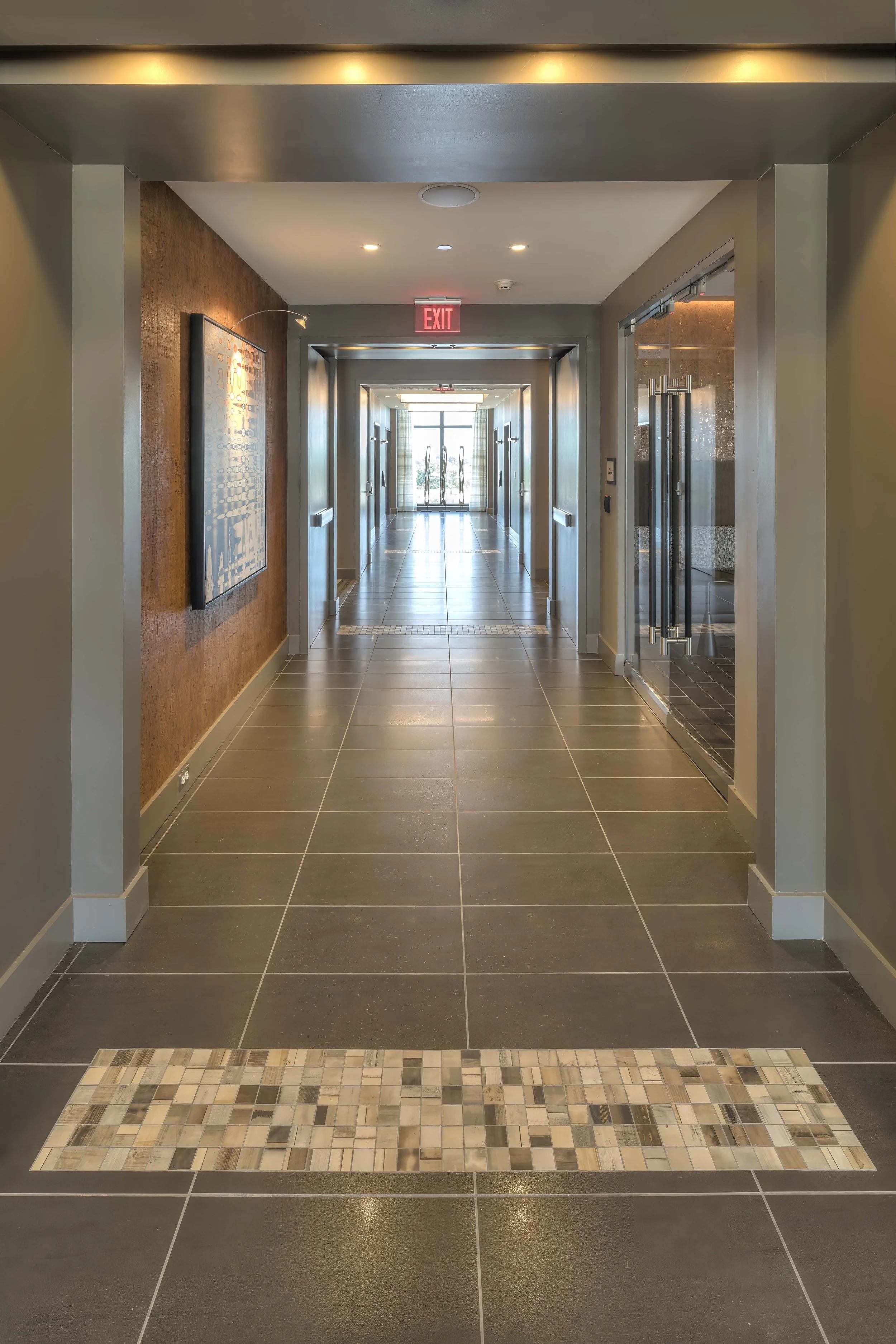 Hotel hallway with tiled floor, artwork on the left wall, glass elevator on the right, and exit doors at the end.