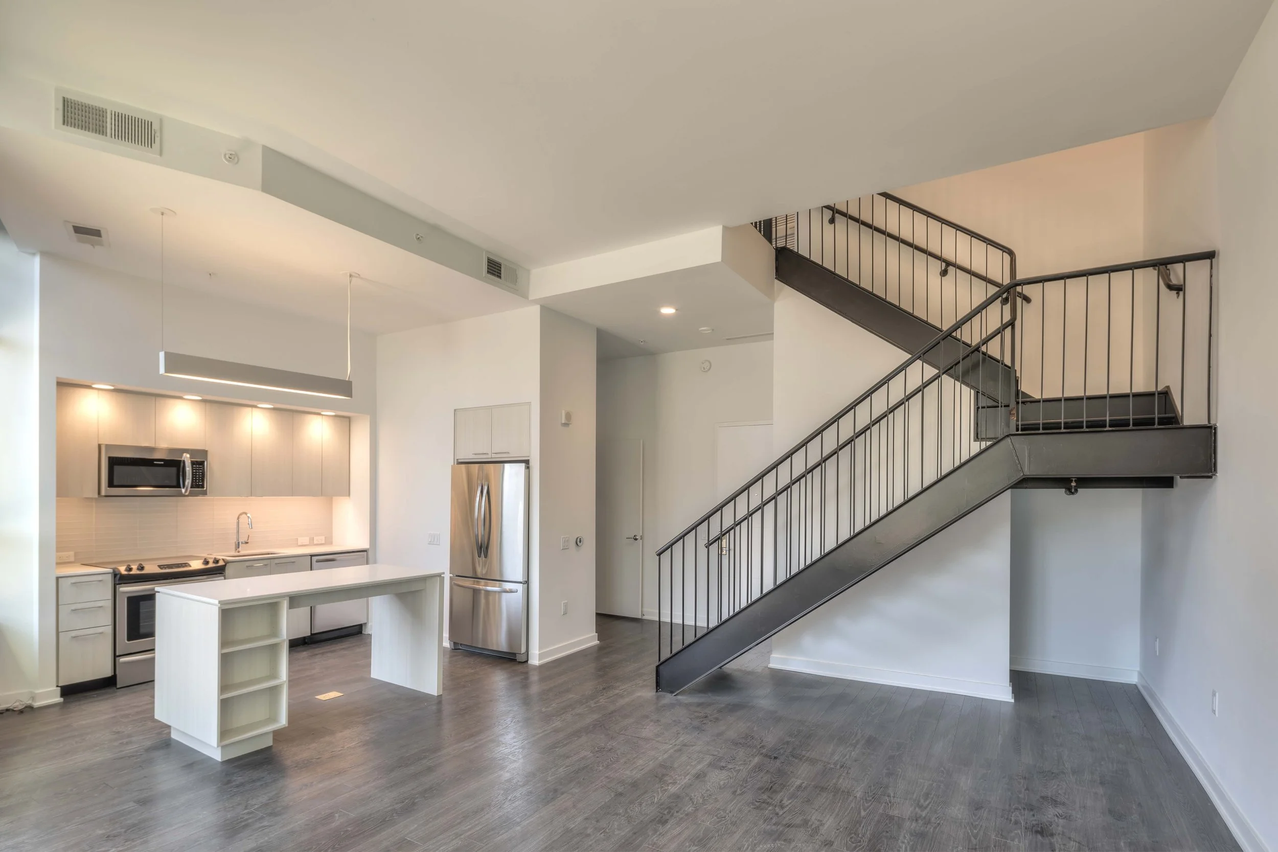 Open-concept kitchen with white cabinets, stainless steel refrigerator, microwave, stove, and a central white island, next to a staircase with black railings in a modern, minimalistic living space.