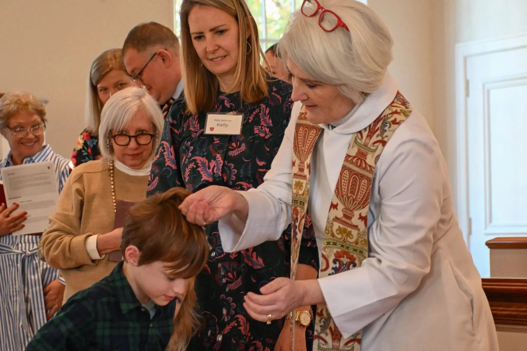 A group of people gathered around a young girl and an older woman, who is wearing glasses on her head, during a baptism or religious ceremony in a well-lit room. The older woman appears to be performing the ritual on the young girl.
