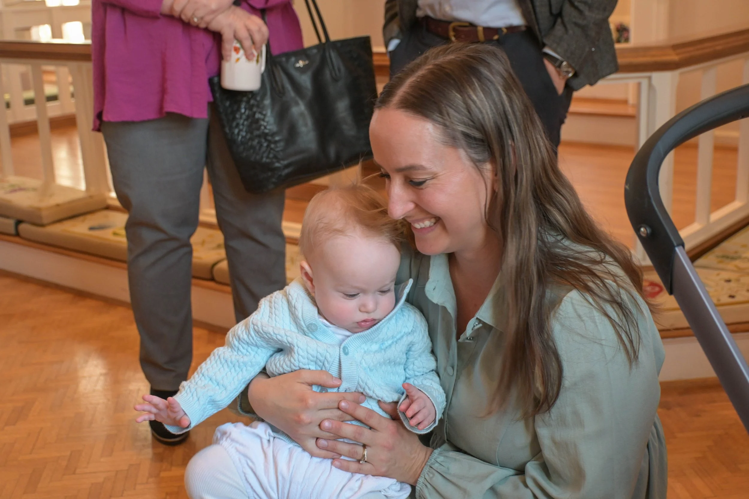A woman with long brown hair holding a young child with red hair in her arms, both smiling and looking at each other, in an indoor setting with wooden floors and a staircase in the background, while two other people stand nearby.