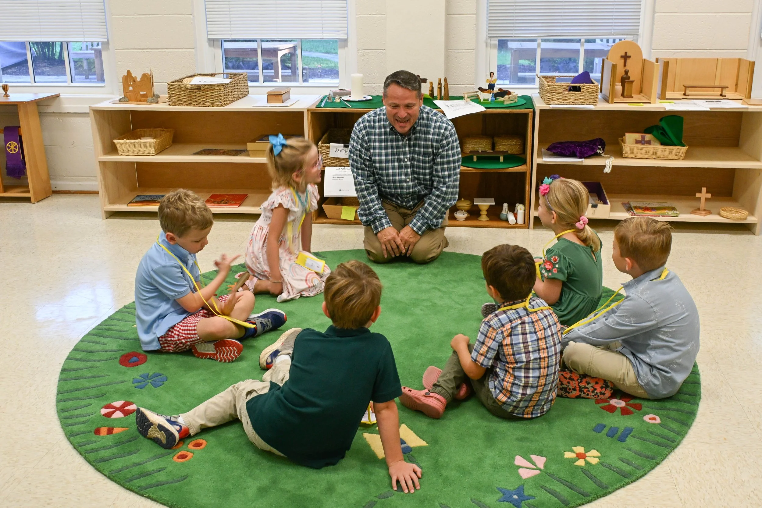 A male teacher kneeling on a green circular rug with children sitting around him, all in a classroom with shelves and windows in the background.