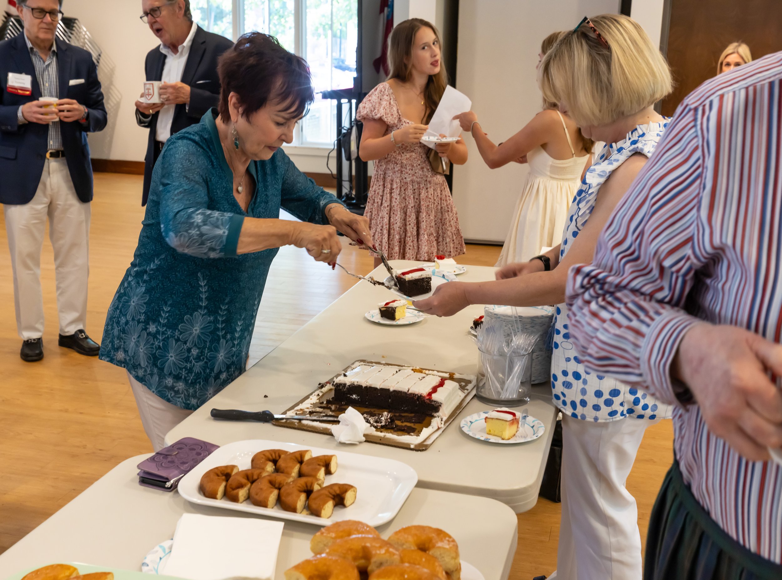 People at a social gathering serving and eating desserts including slices of cake, donuts, and pastries at a buffet table.