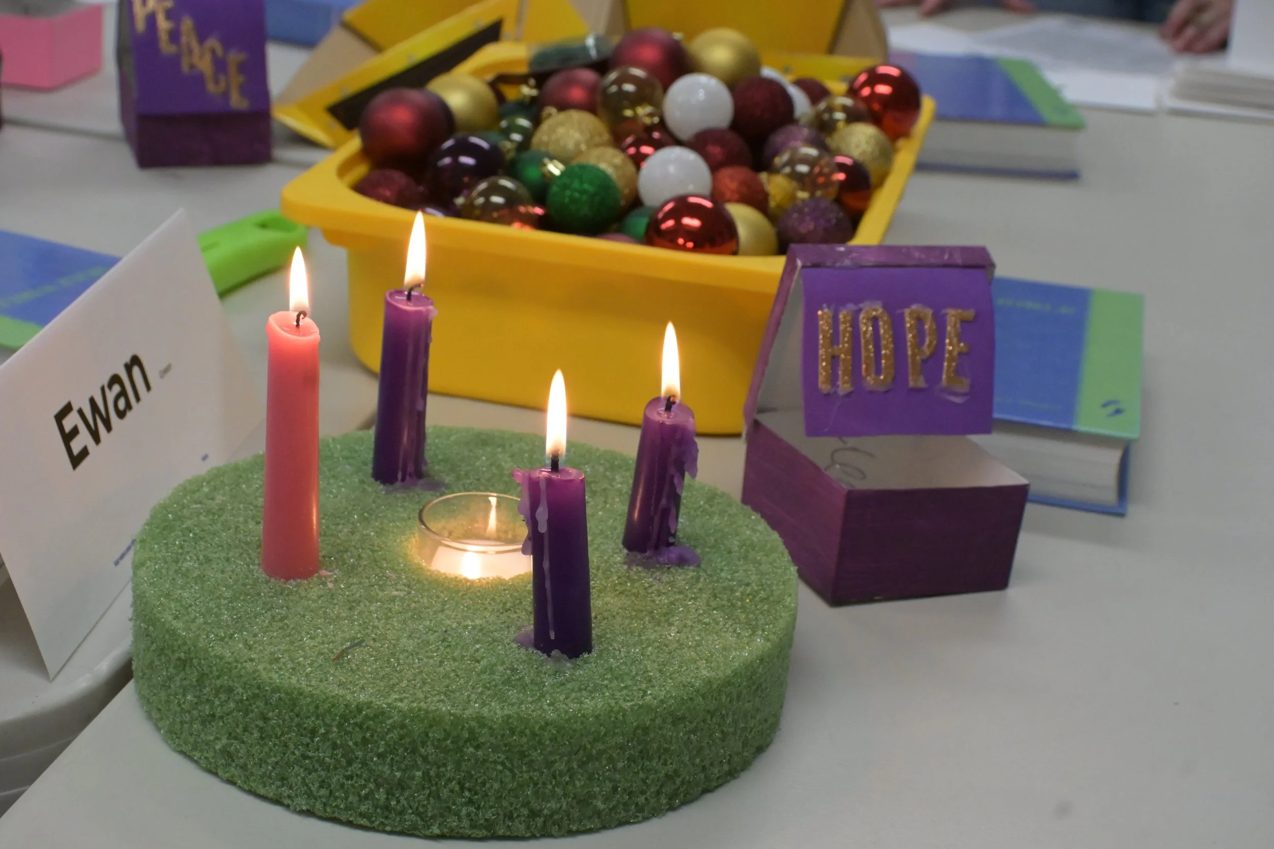 Colorful Christmas-themed table display with a small green candle holder with four lit candles, a yellow container filled with assorted Christmas ornaments including red, gold, white, and purple baubles, and motivational signs with the words 'Hope' and a name tag reading 'Ewan'.