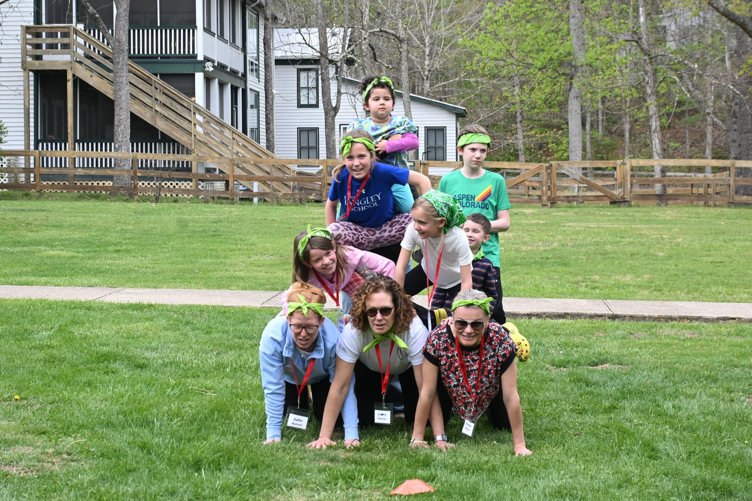 A group of children and adults forming a human tunnel outdoors on a grassy area, with a house and trees in the background.