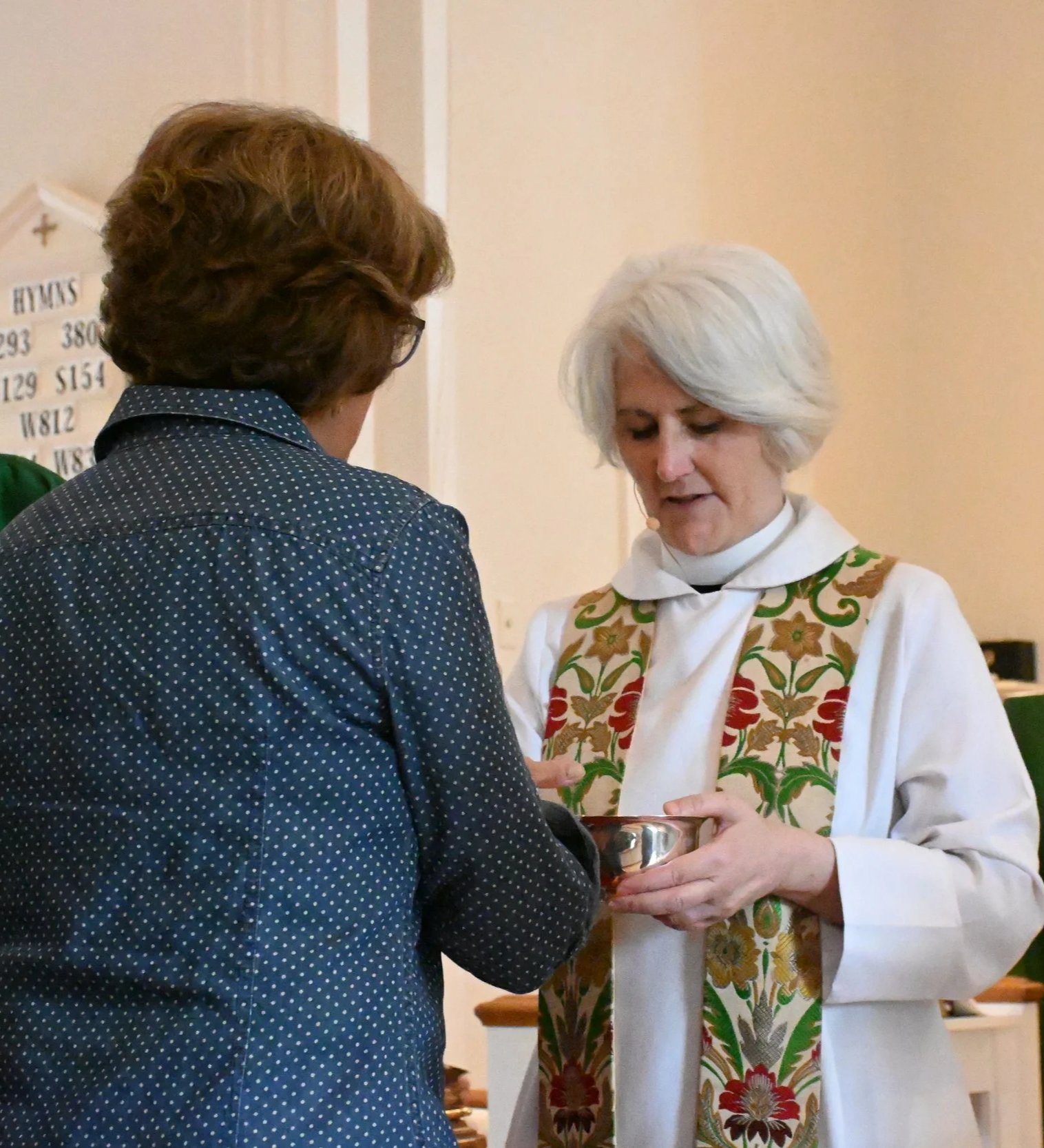 A woman with short white hair wearing a white robe and a colorful embroidered stole is holding a silver dish. She is standing in front of another woman with curly brown hair and glasses, wearing a dark blue polka-dot shirt, who appears to be receivin
