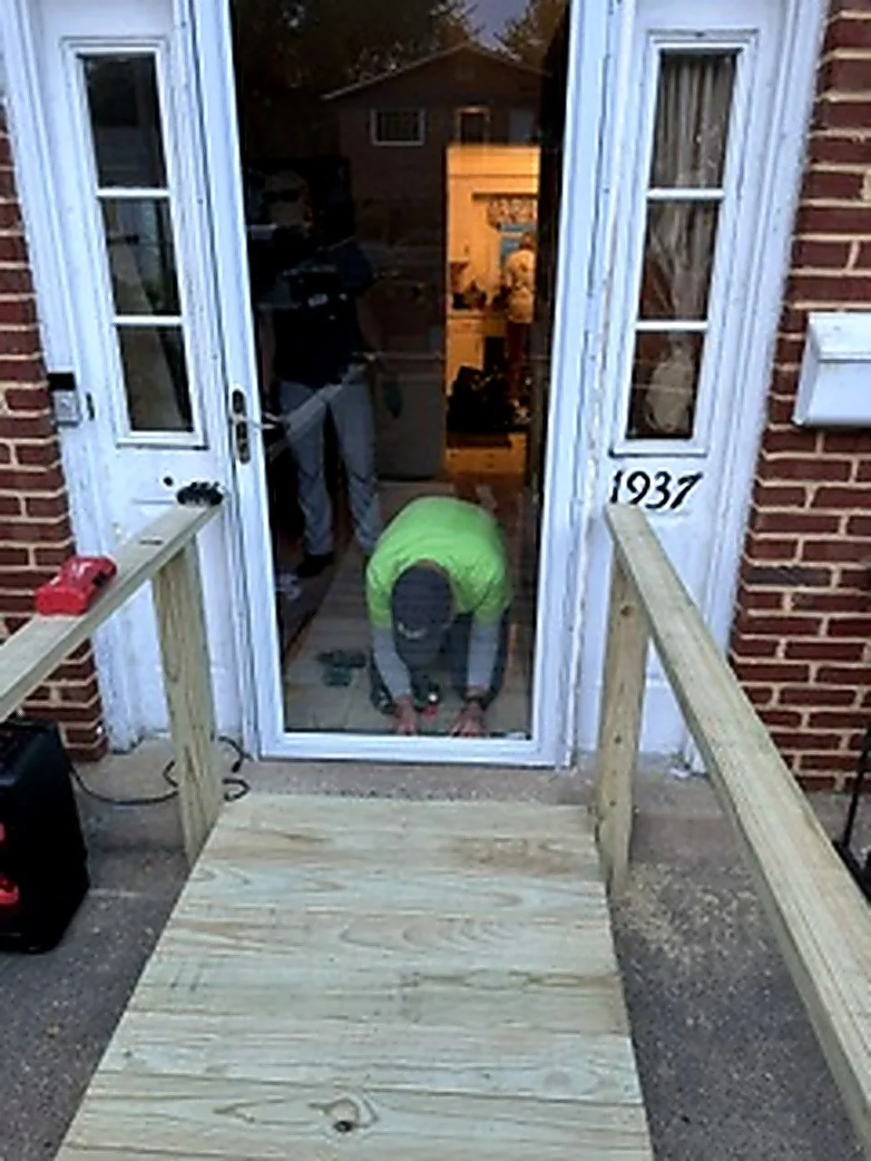 Person working on constructing a wooden ramp at the front door of a house with brick walls, glass door, and house number 1937.