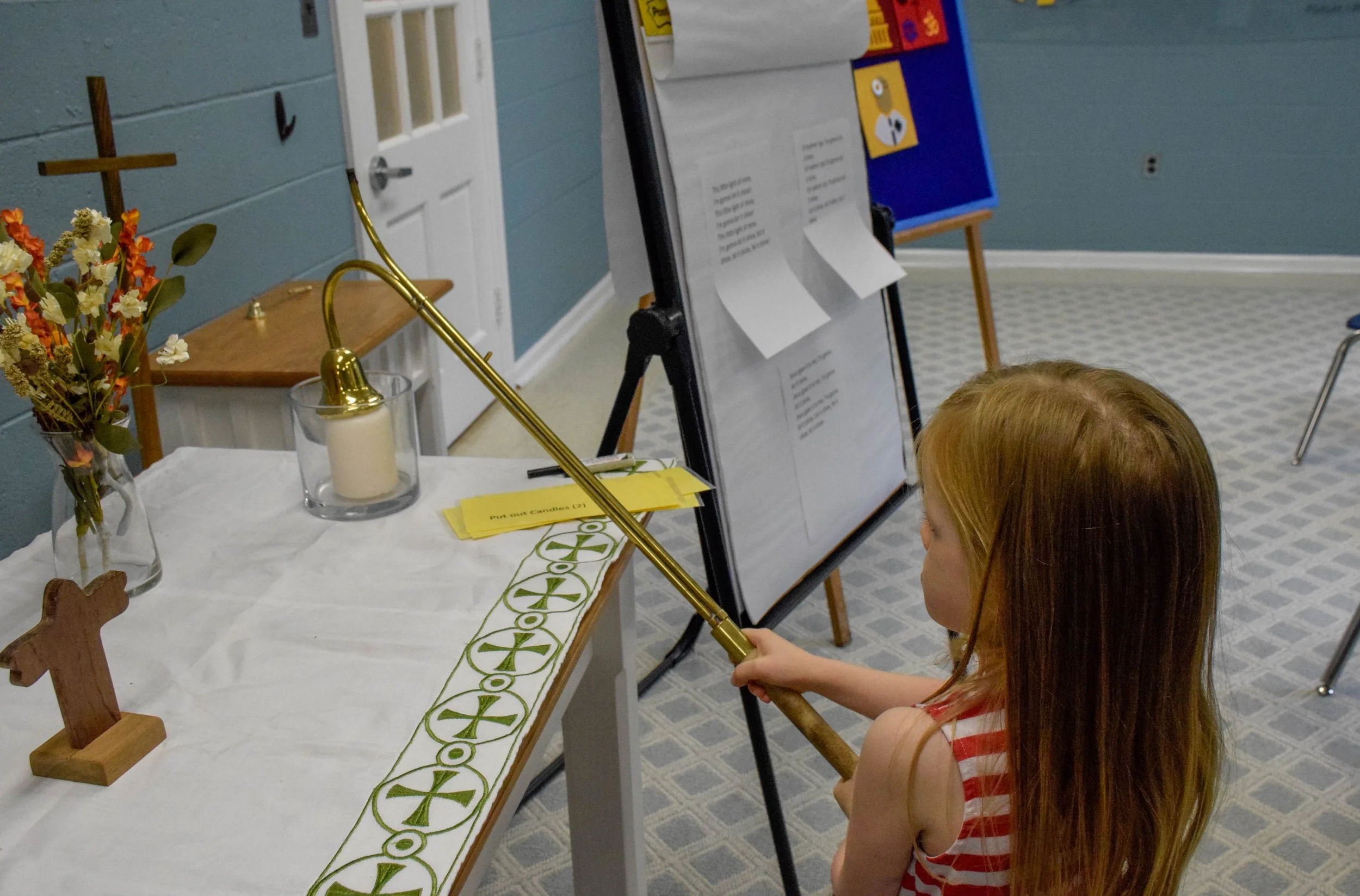 A young girl with red hair and a red and white striped sleeveless top holding a golden sword, looking at a religious setup on a table with a wooden cross, flowers in a vase, a lit candle, and a cloth with green symbols. There is a stand with papers and a door in the background.