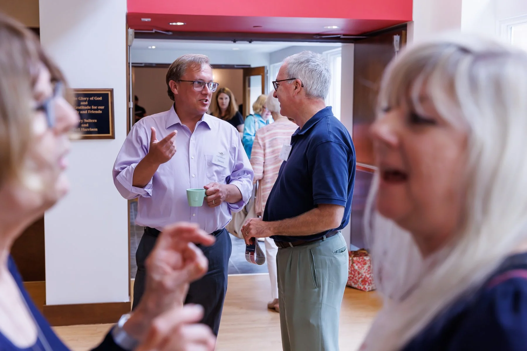 Group of middle-aged people engaging in conversations at a social gathering indoors, some holding coffee mugs.