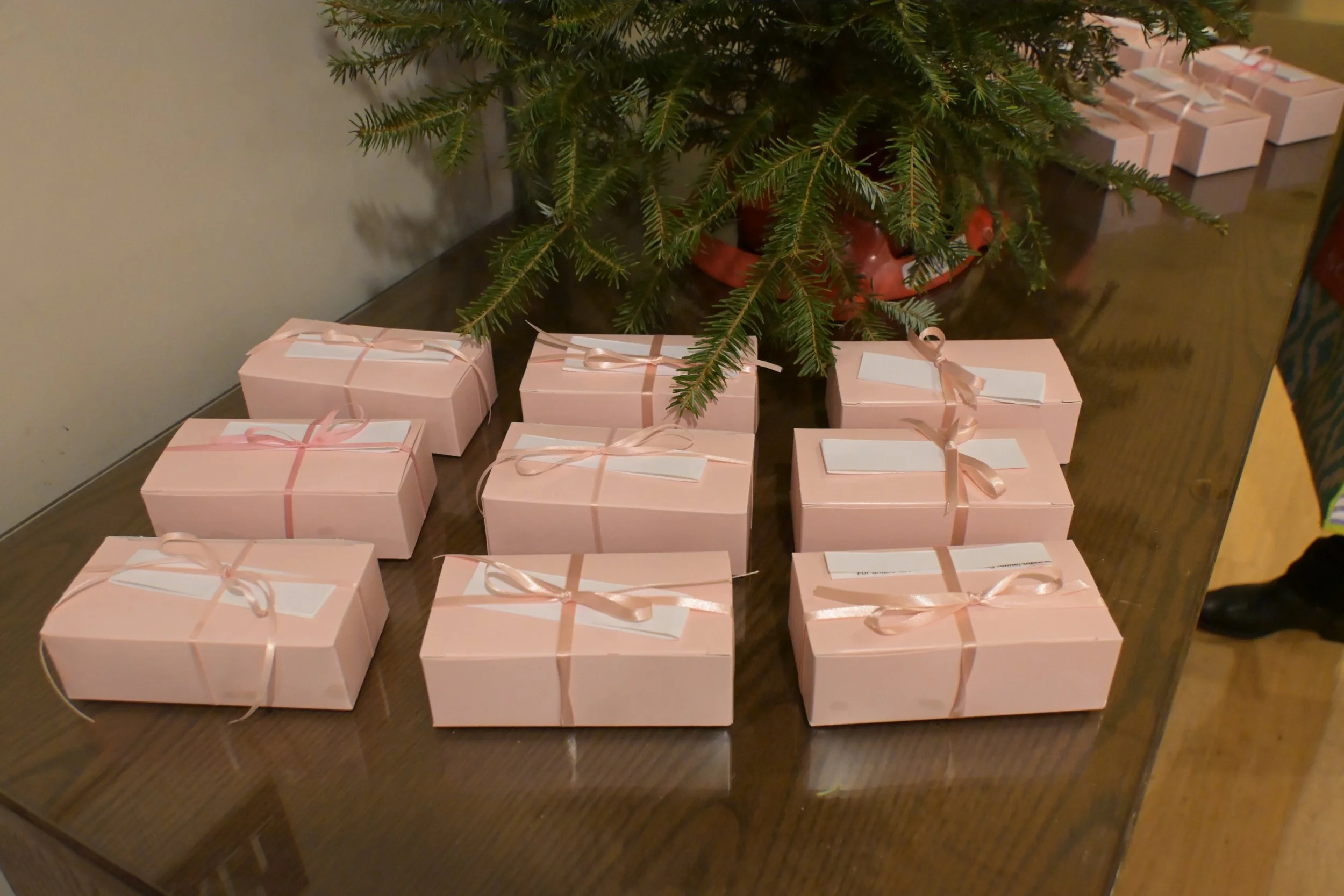 Pink gift boxes tied with pink ribbons arranged on a wooden table beneath a Christmas tree.