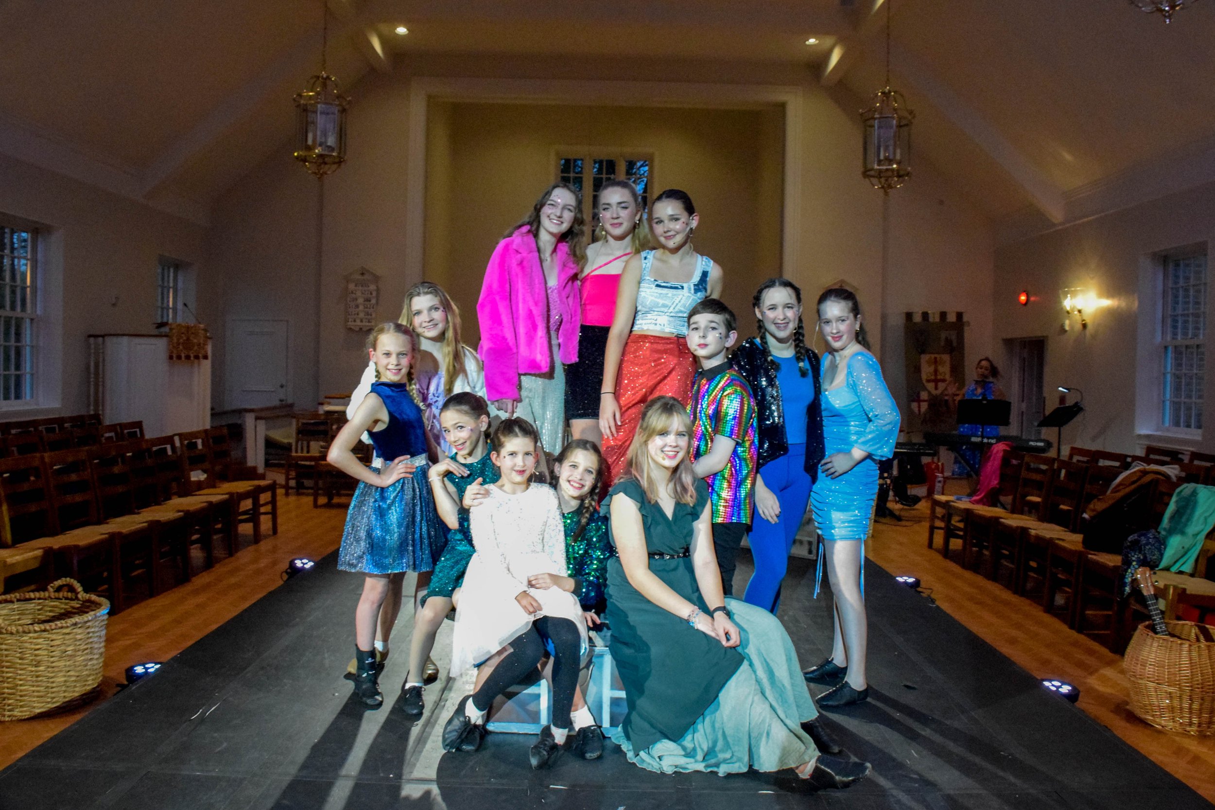 Group of children and young teens in colorful costumes posing on a stage in a hall with wooden floors and empty chairs along the sides.