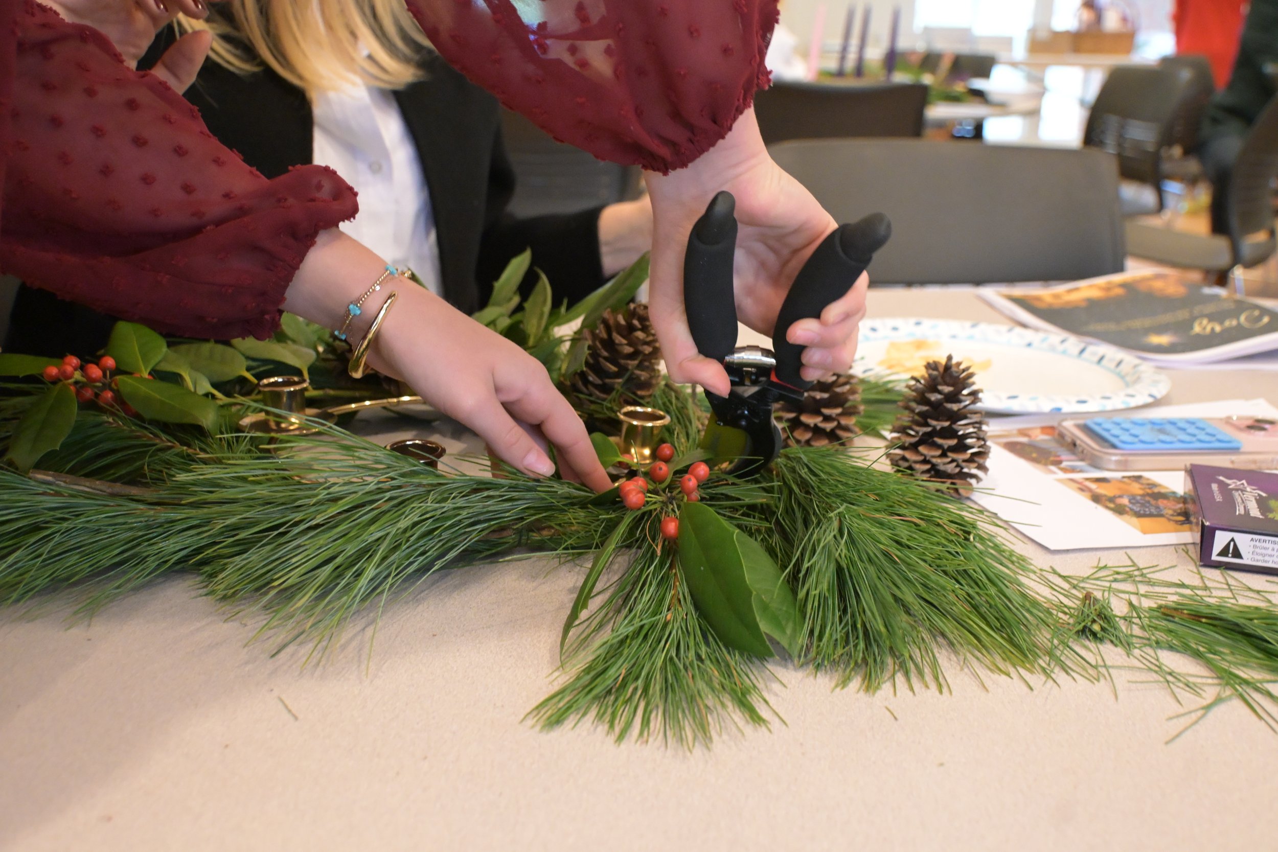 Person arranging festive greenery, including pine branches, holly leaves, pinecones, and red berries, on a table for decoration.