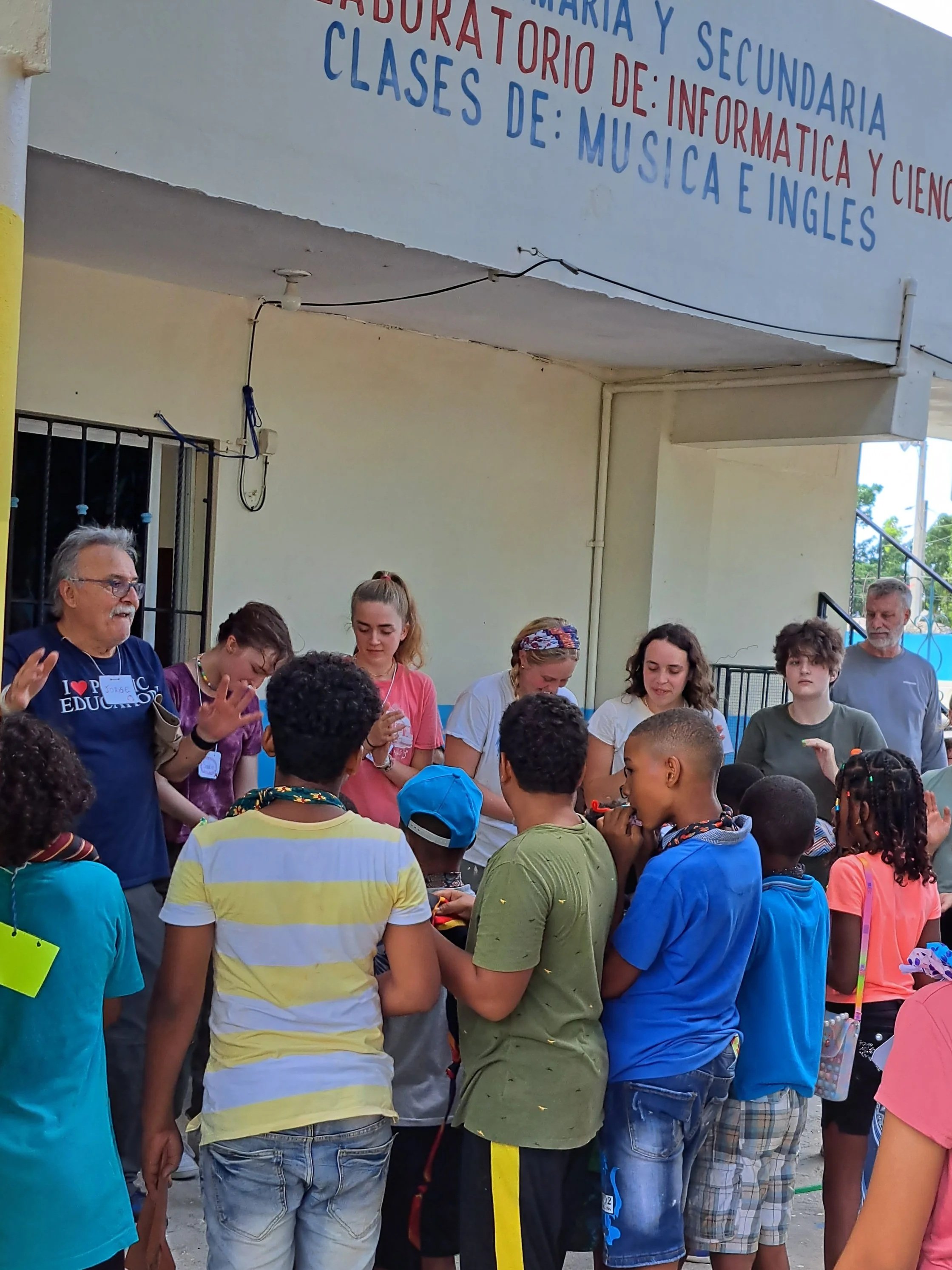 Children gathered around teachers or facilitators outside a school building, participating in an activity, with a sign indicating the school offers classes in computer science, music, and English for high school students.