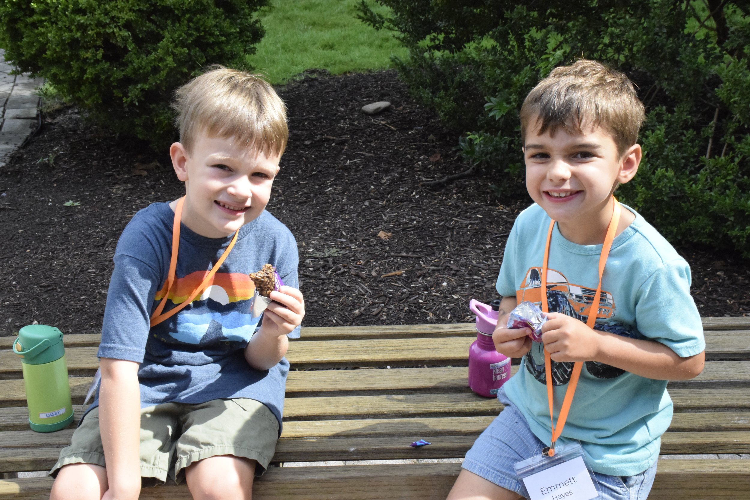 Two young boys sitting on a wooden bench outdoors, smiling and holding snacks, with water bottles next to them and lush greenery in the background.