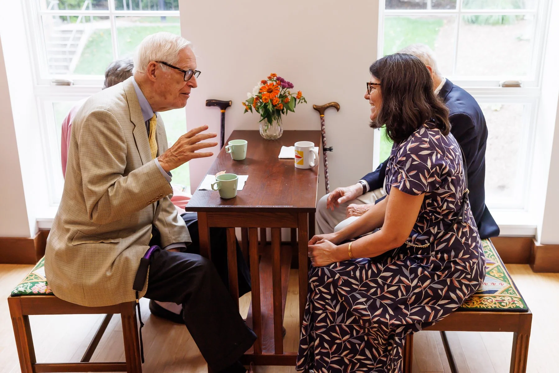 An elderly man and a woman are sitting across from each other at a wooden table, engaged in a conversation. The man is gesturing with his hand, wearing glasses, a beige blazer, and a yellow tie. The woman is smiling, wearing glasses, a patterned dress, and gold jewelry. On the table are three mugs and a flower vase with orange and purple flowers. Two additional people are partially visible in the background, and two walking canes are resting against the wall behind the table. Large windows let in natural light.