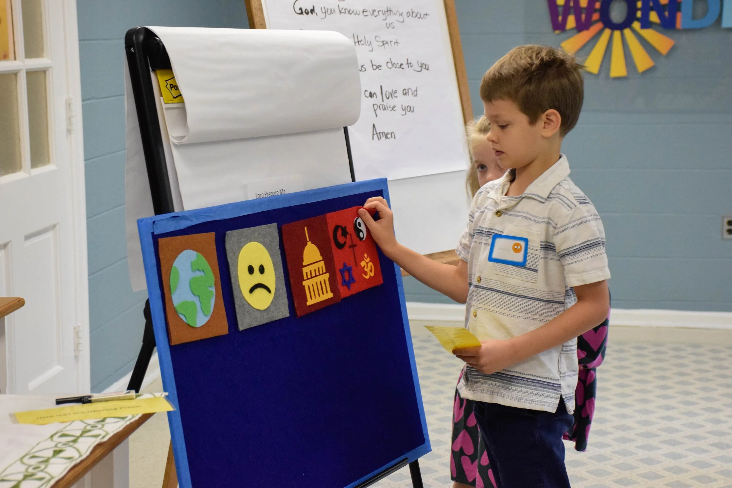 Two children, a young boy and girl, engaging in an activity at a classroom or church event. The boy is standing near a blue display board with felt icons depicting the earth, a sad face, the Capitol, a star of David, and a peace sign. The girl is partially visible behind the boy, watching him. In the background, a whiteboard with handwritten text and a colorful decoration on the wall are visible.