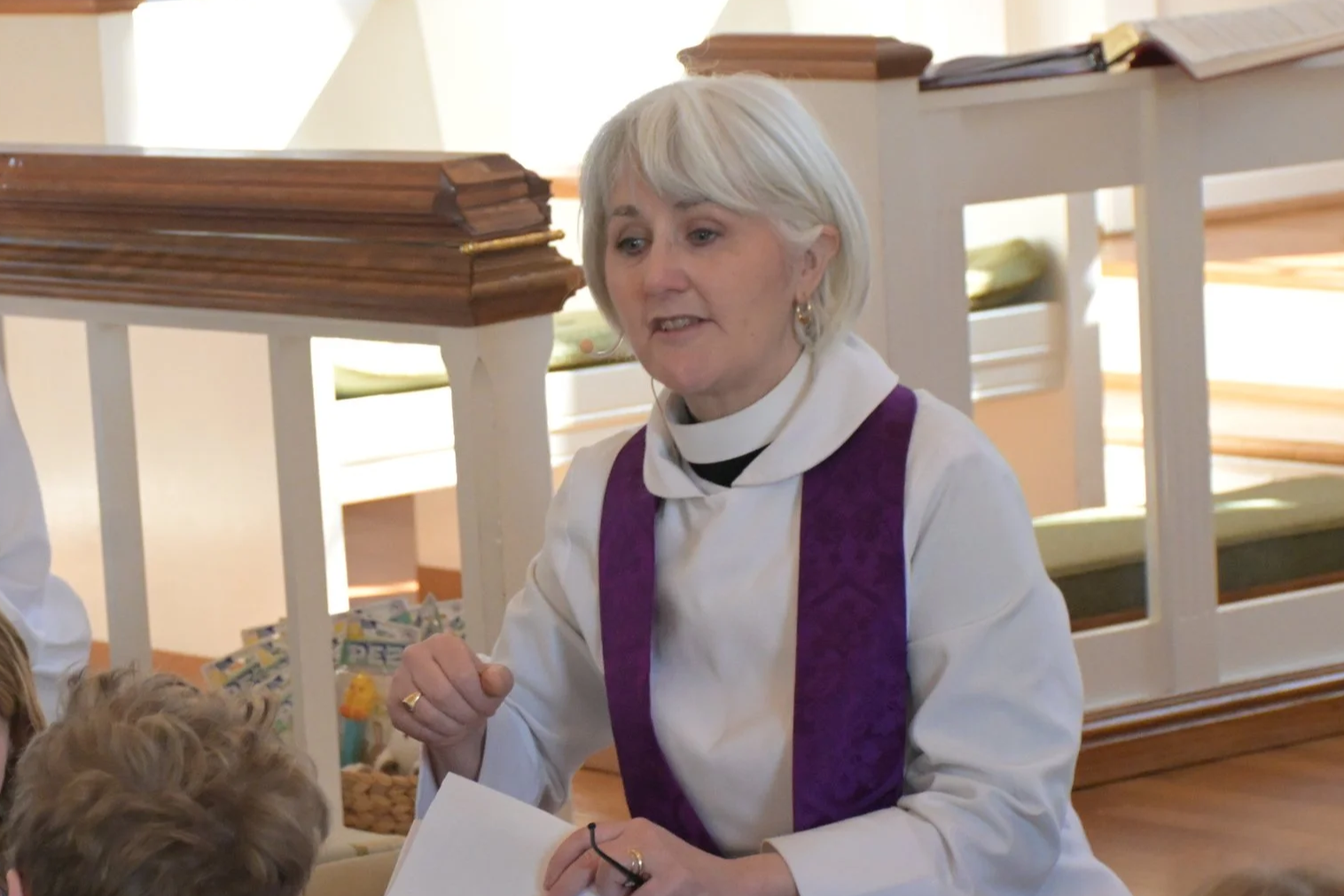 An older woman with short, light gray hair, wearing clerical attire including a white garment and a purple stole, speaking to a group of children inside a church or chapel.