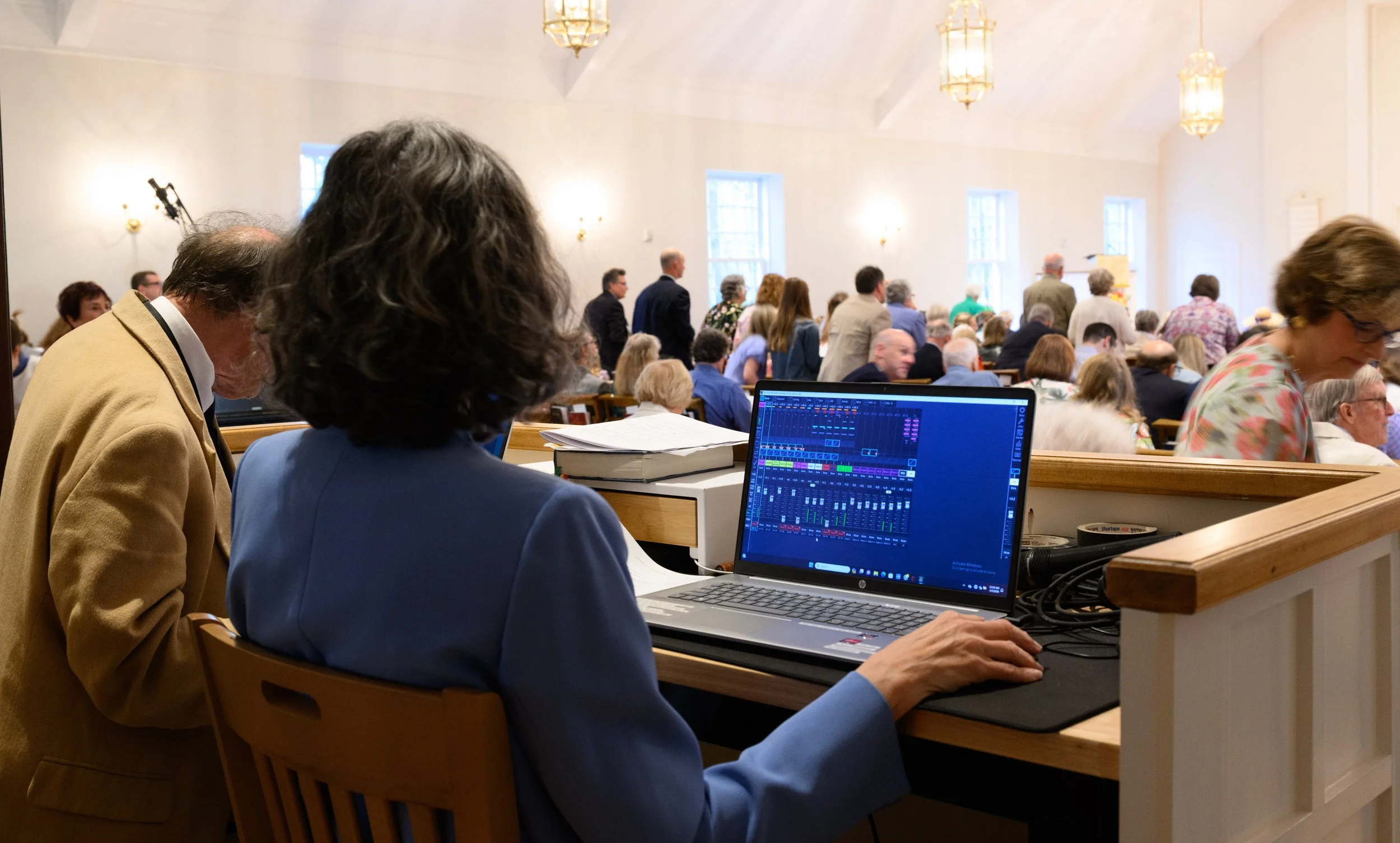 People attending a gathering or conference in a bright room with large windows and hanging lights. In the foreground, a person in a blue suit is working on a laptop displaying audio or music software. Other attendees are seated or standing, some engaged in conversations.