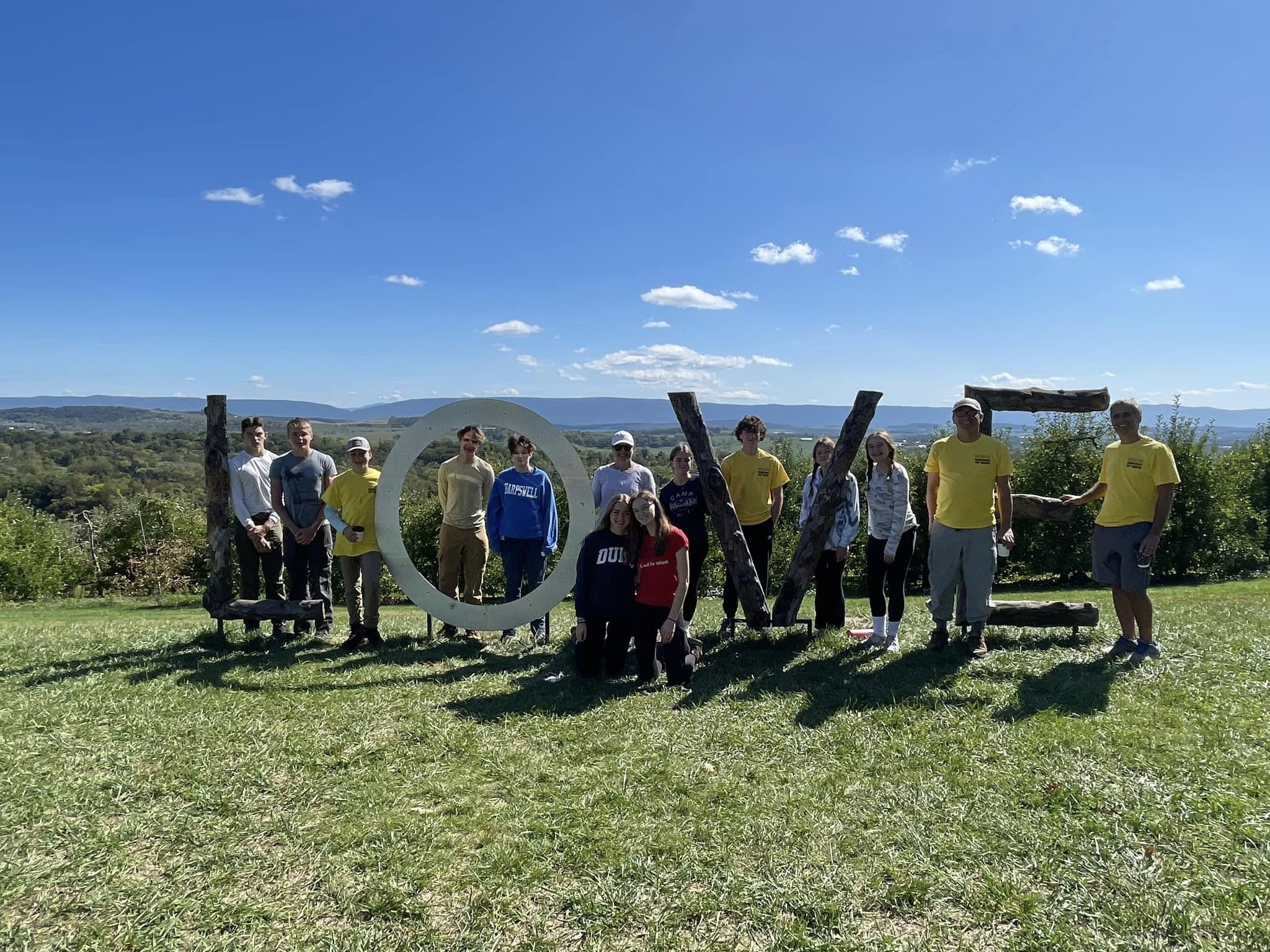 Group of people standing outdoors on grass with the word 'LOVE' spelled out using wooden logs and metal circle, under a blue sky with clouds.