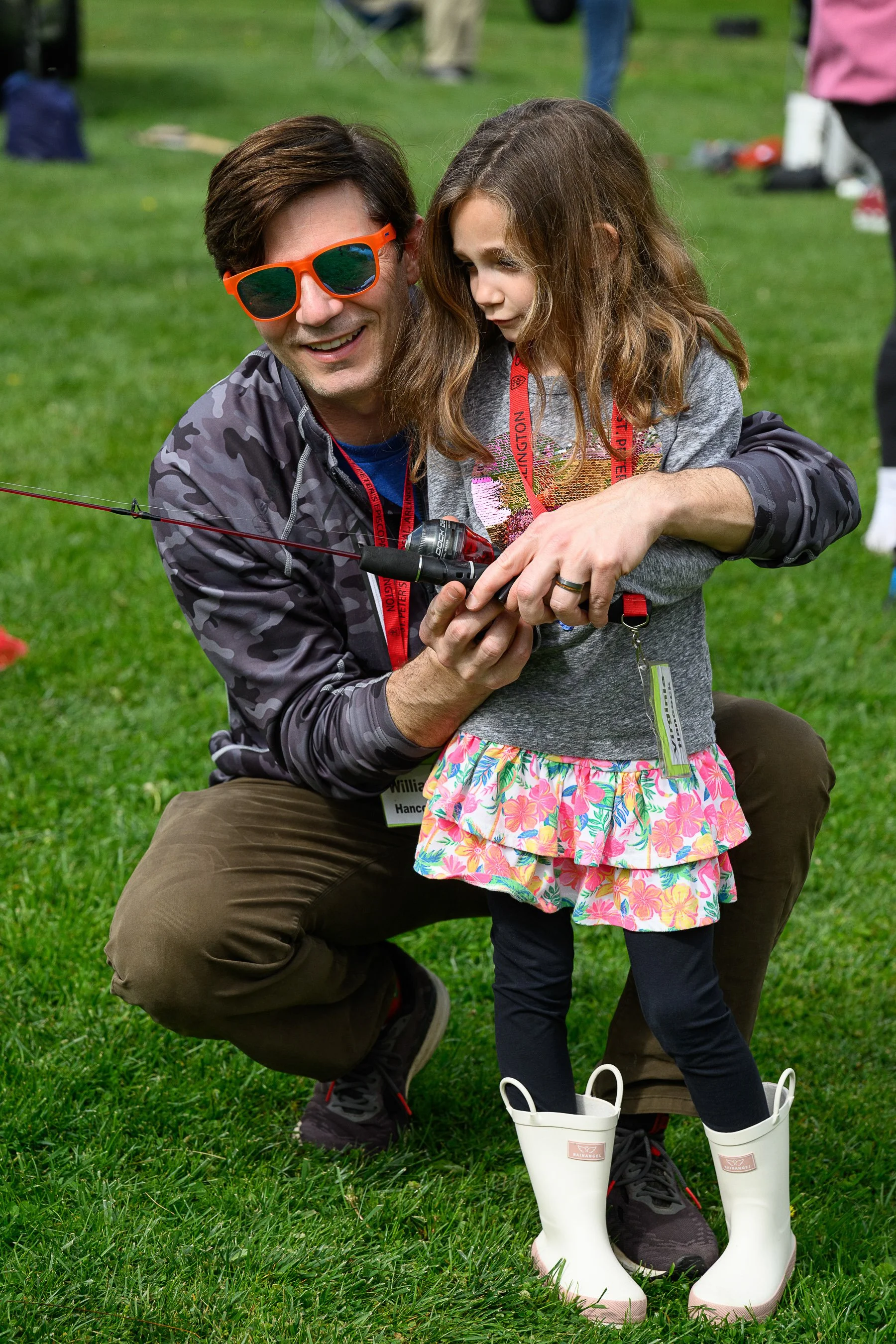 A man and a young girl in outdoor clothing, with the man showing her how to use a fishing rod on a grassy field during an outdoor event.
