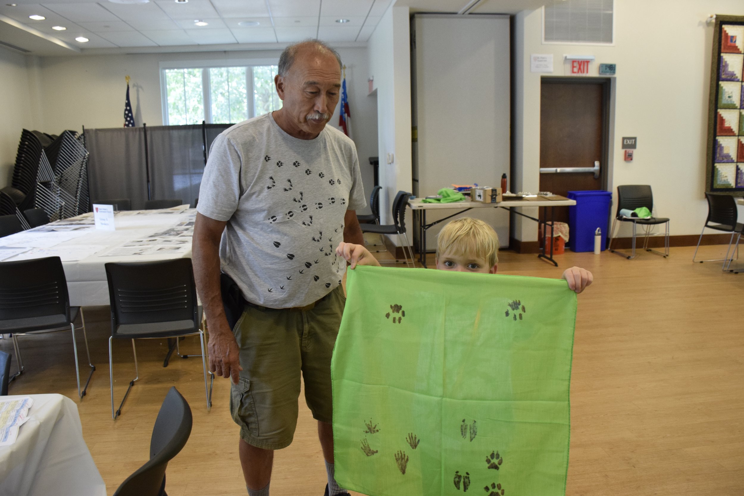An older man with a mustache and a young boy with blonde hair holding a green cloth with black footprints printed on it in a community room.
